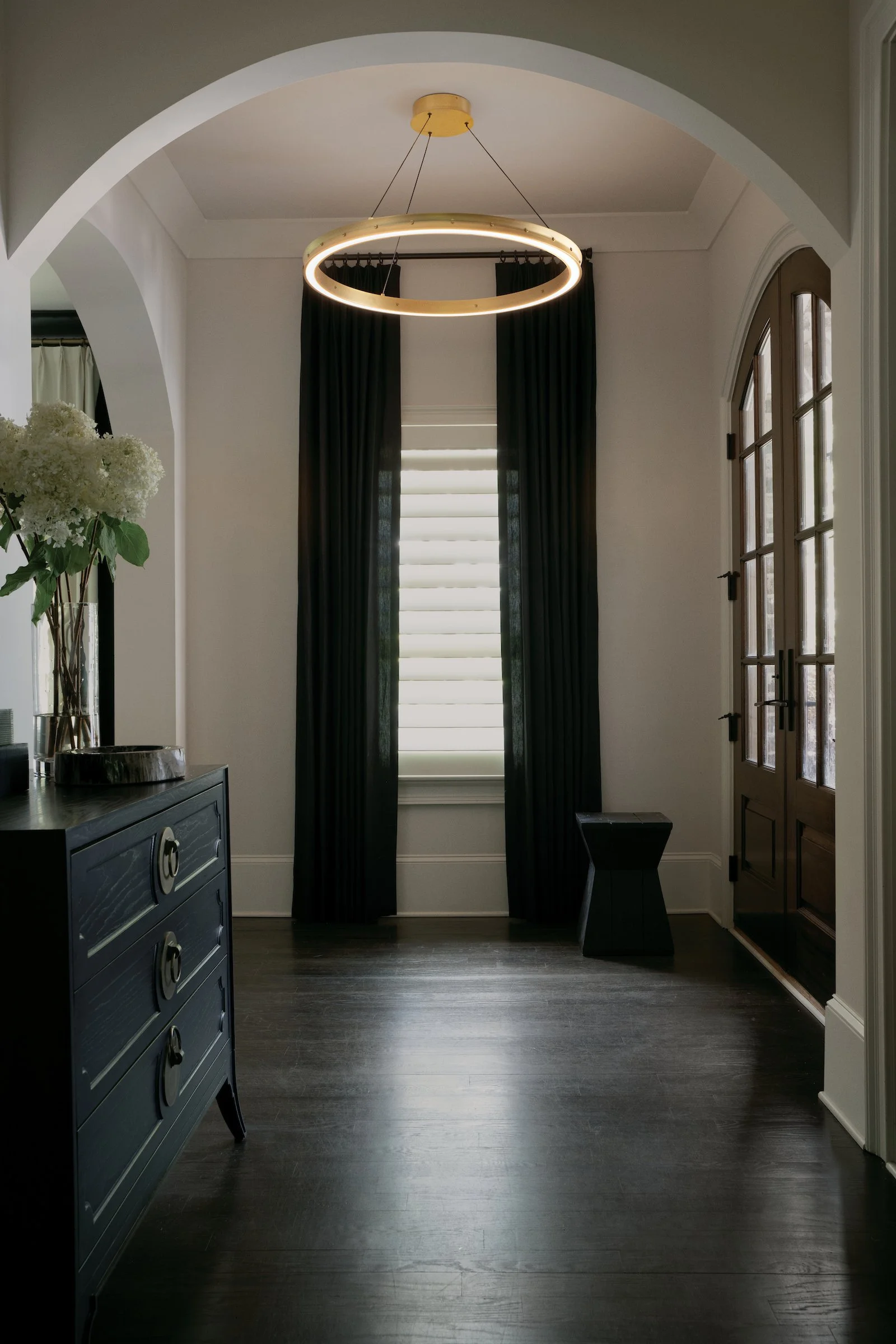 Interior view of a room with a modern circular chandelier hanging from the ceiling, dark curtains covering the window, a dark wood floor, a black side table, and a dark dresser with a glass vase of white flowers.