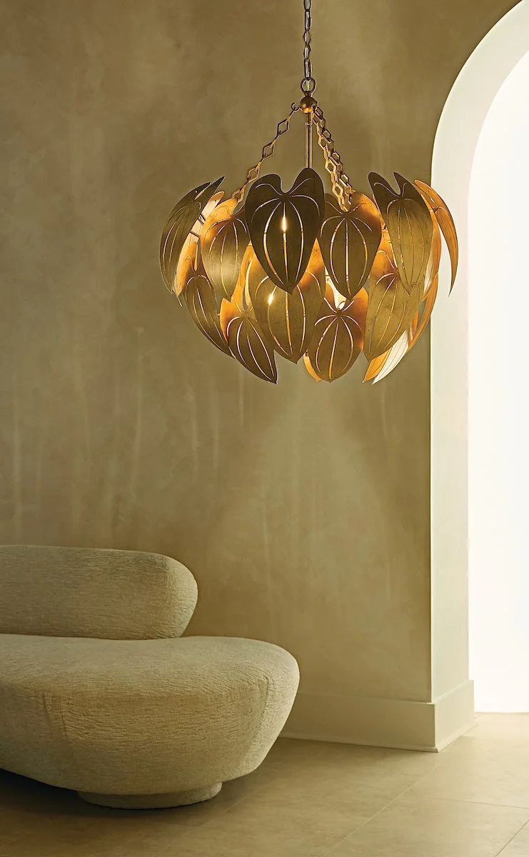 A decorative chandelier made of overlapping metallic leaf-shaped panels hanging from the ceiling in a modern interior space, with a beige sofa to the left and a wall with an archway to the right.