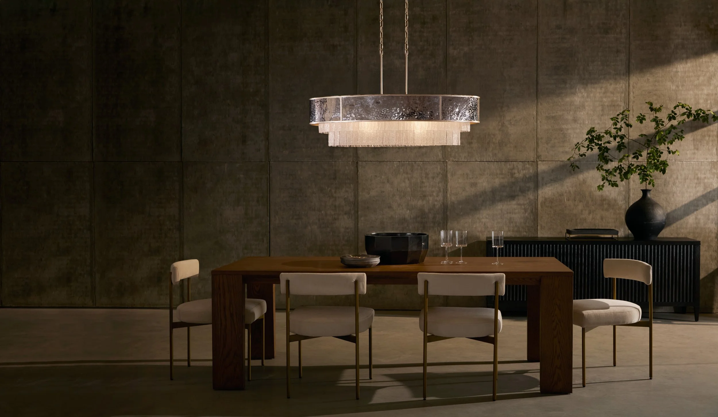 Modern dining room with a wooden table, six white chairs, a large decorative chandelier, a black vase with green plant, black bowl, and two glasses on the table, against a textured gray wall.