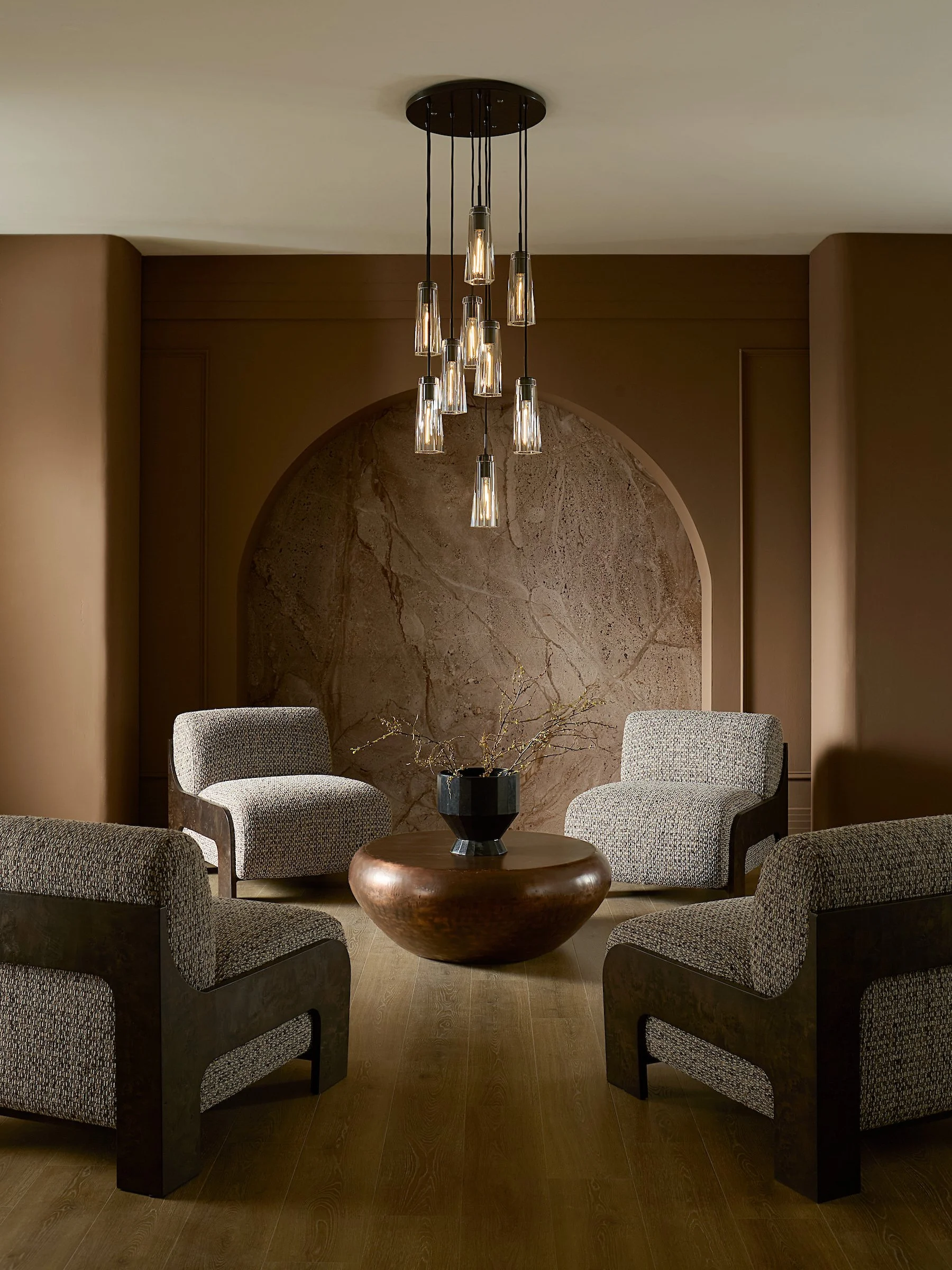 A modern living room with a round wooden table, six upholstered armchairs with patterned fabric, a black vase with thin branches, a brown textured wall, and a hanging chandelier with multiple exposed bulbs.
