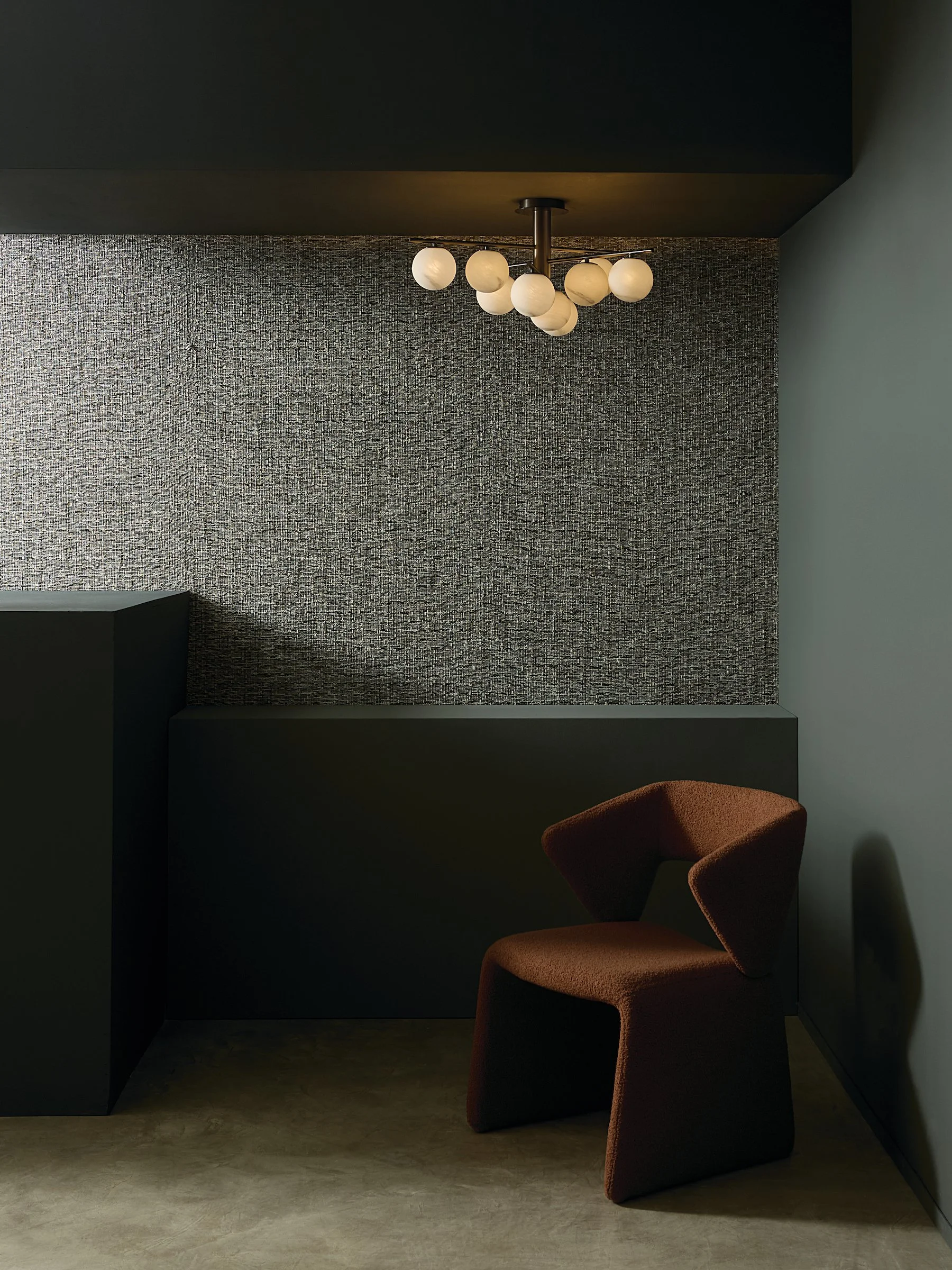 Corner of a room with a hanging ceiling light, an orange upholstered chair, a textured wall with gray and black patterns, and a light gray wall.