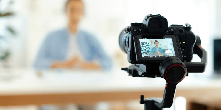 A camera on a tripod capturing an interview with a person sitting at a desk, blurred background.