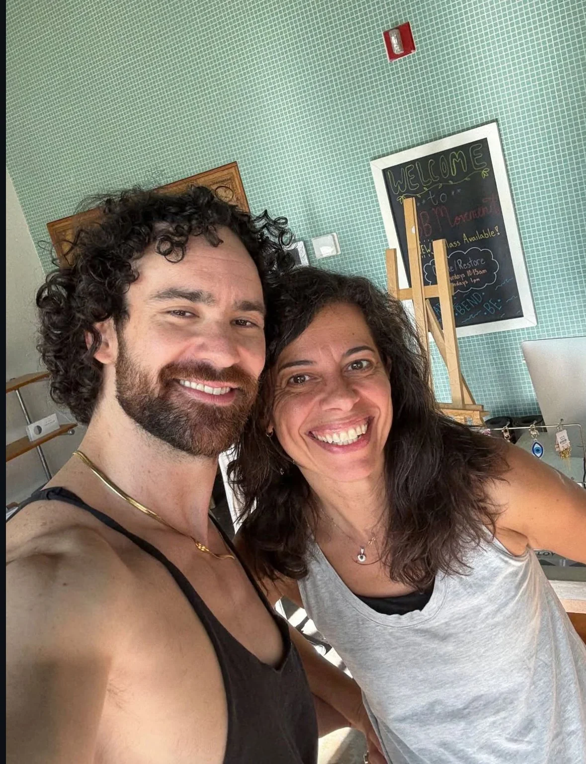 Two people taking a selfie indoors, smiling, in front of a chalkboard with a welcome message and a jewelry display in the background.
