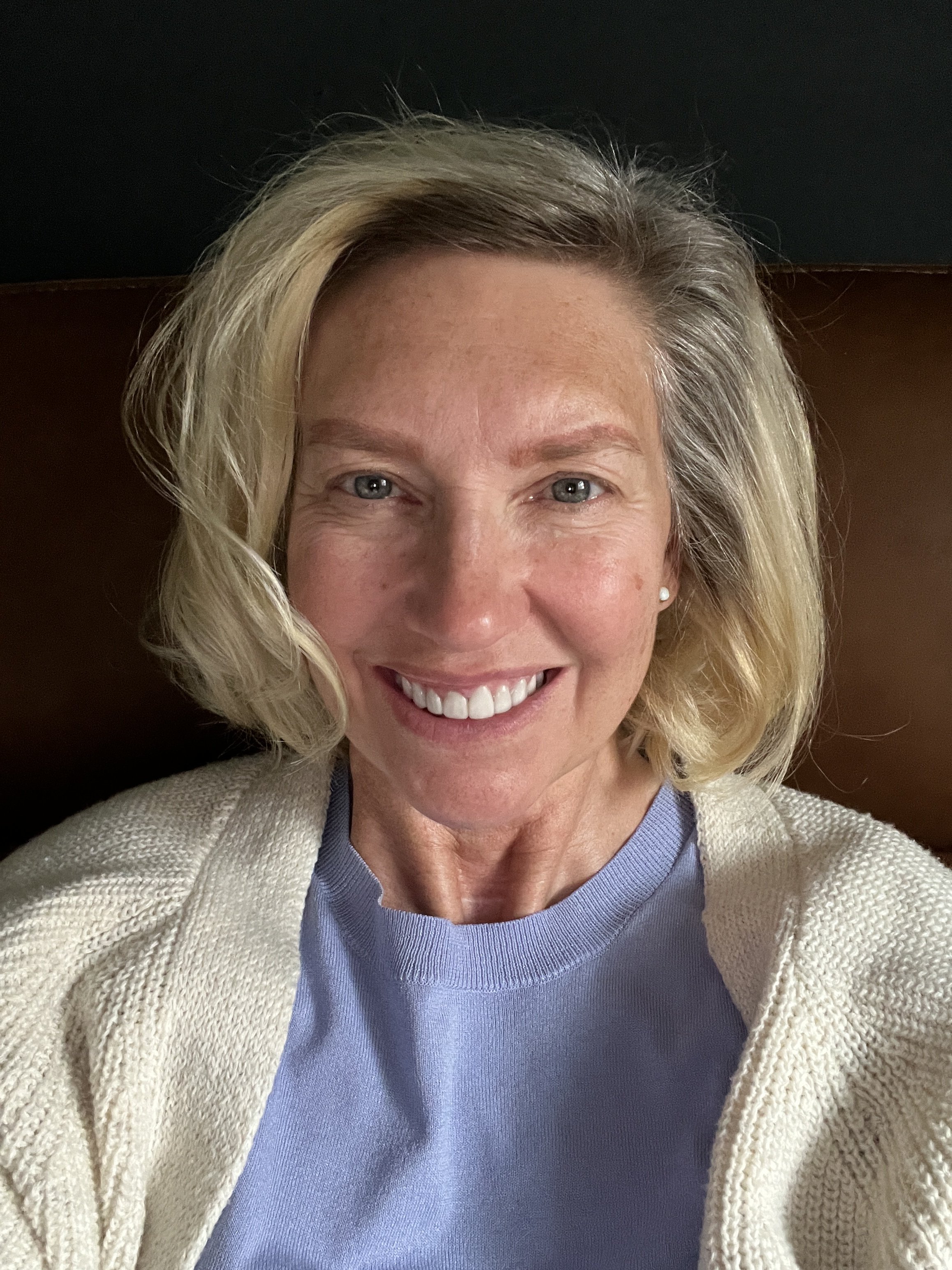 Close-up of a smiling woman with blonde hair, blue eyes, and a light complexion, wearing a cream-colored cardigan over a blue sweater.