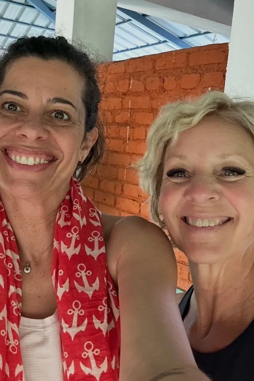 Two women smiling and taking a selfie indoors, with a brick wall and a window with a metal roof in the background.