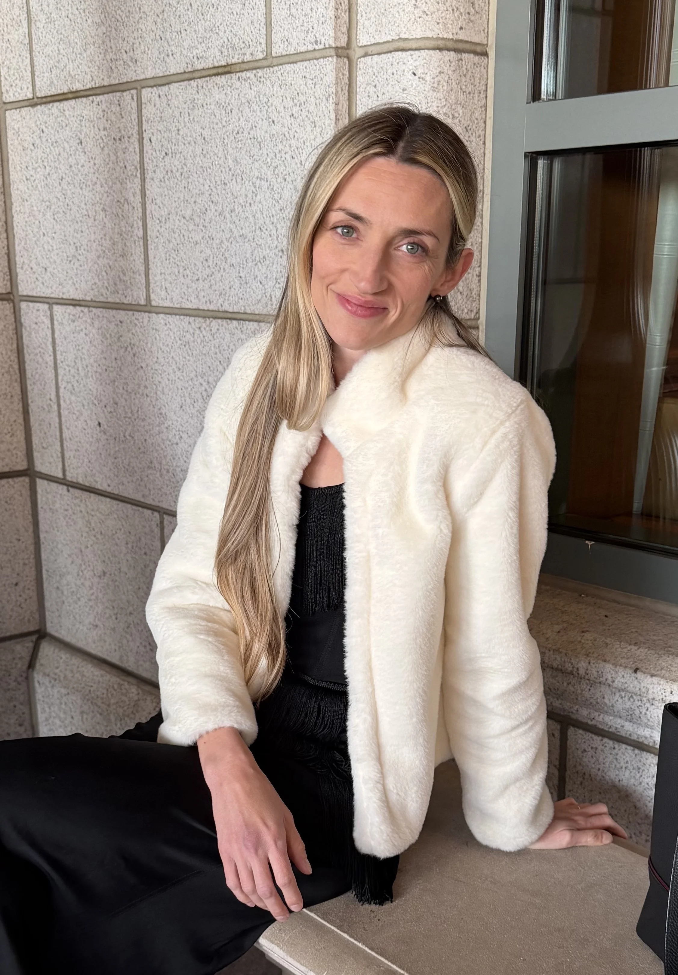A woman with long blonde hair and blue eyes smiling while sitting on a porch, wearing a cream-colored coat and a black dress.