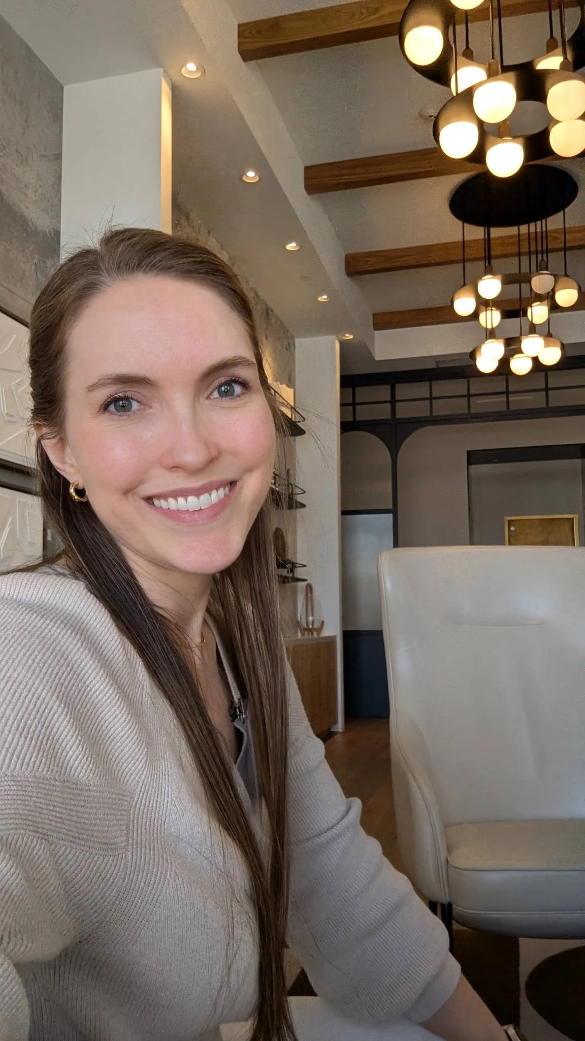 A woman with brown hair and blue eyes smiling in a modern room with wooden beams, unique light fixtures, and beige chairs.