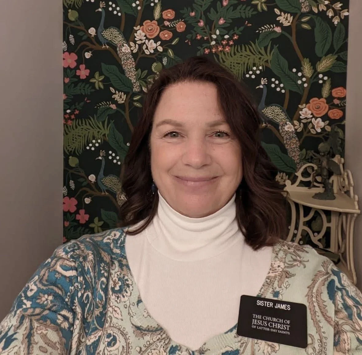 A woman with shoulder-length brown hair wearing a white turtleneck and a patterned cardigan taking a selfie in front of a floral tapestry with peacocks. She is smiling and wearing a name tag that reads 'Sister James, The Church of Jesus Christ of Latter-day Saints.' Behind her is a decorative shelf with small figurines.