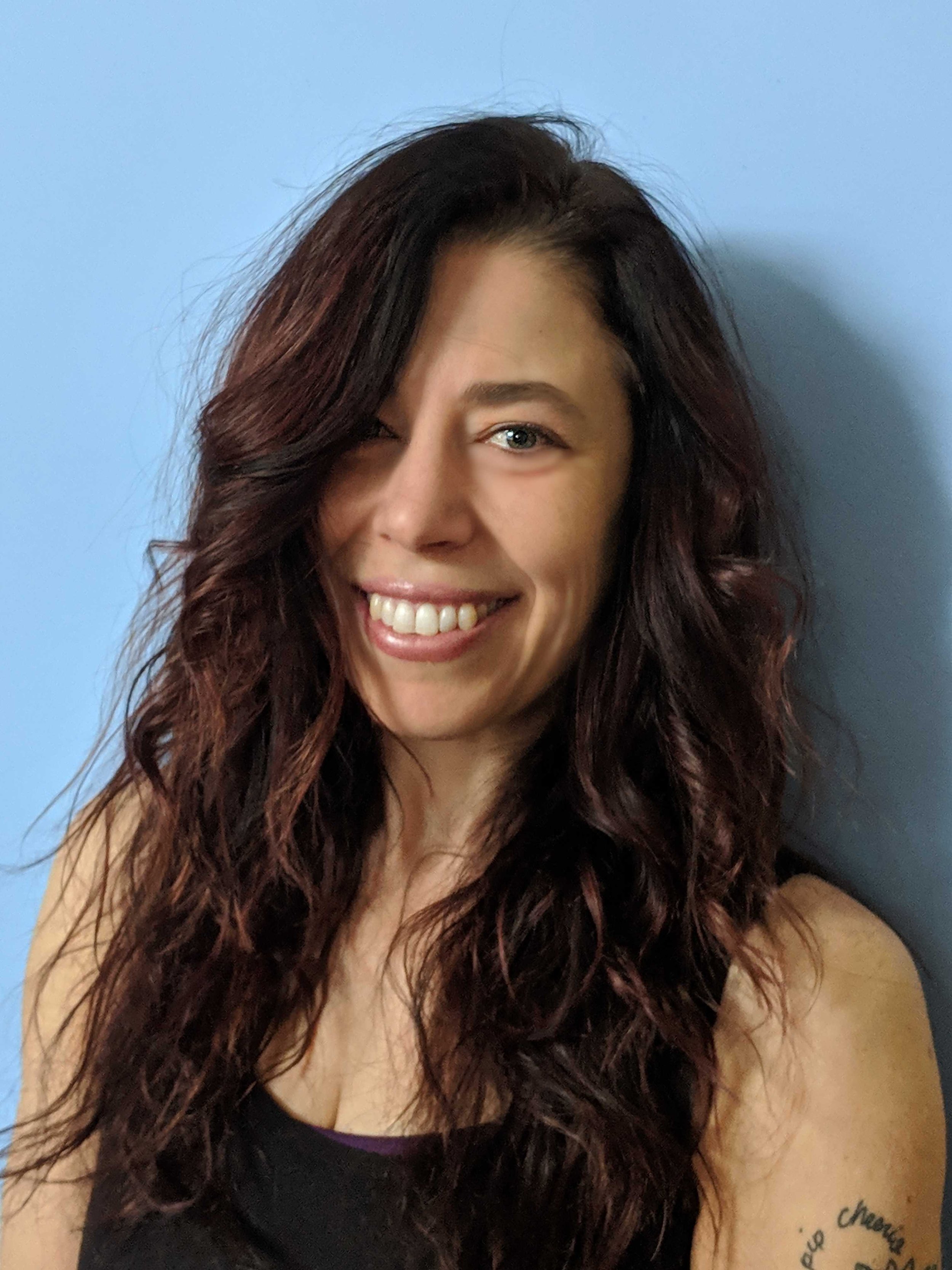 A woman with long, wavy, reddish-brown hair smiling, against a blue wall.