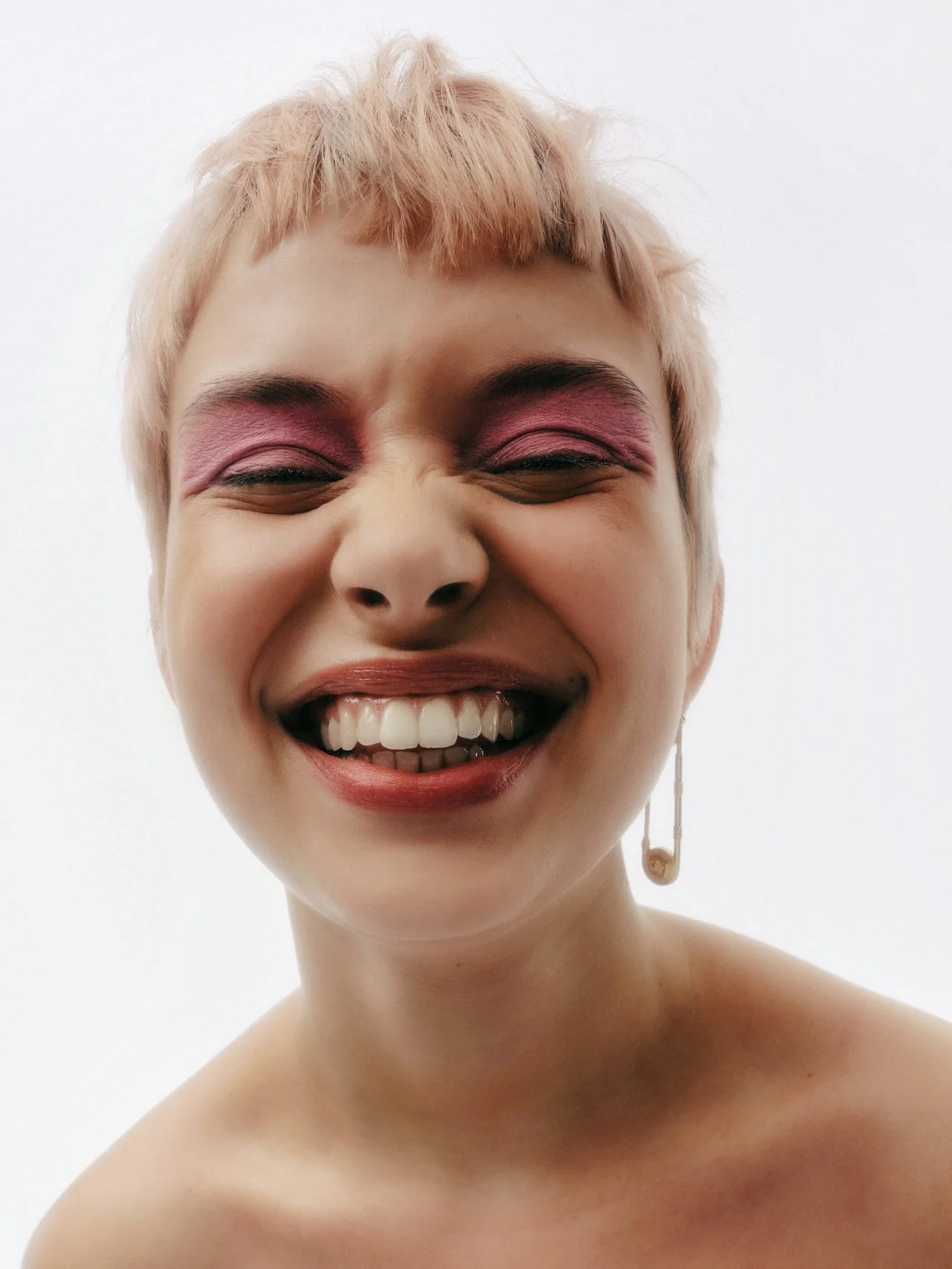 A woman with short, light-colored hair and pink eyeshadow is smiling broadly, showing her teeth, against a plain white background.