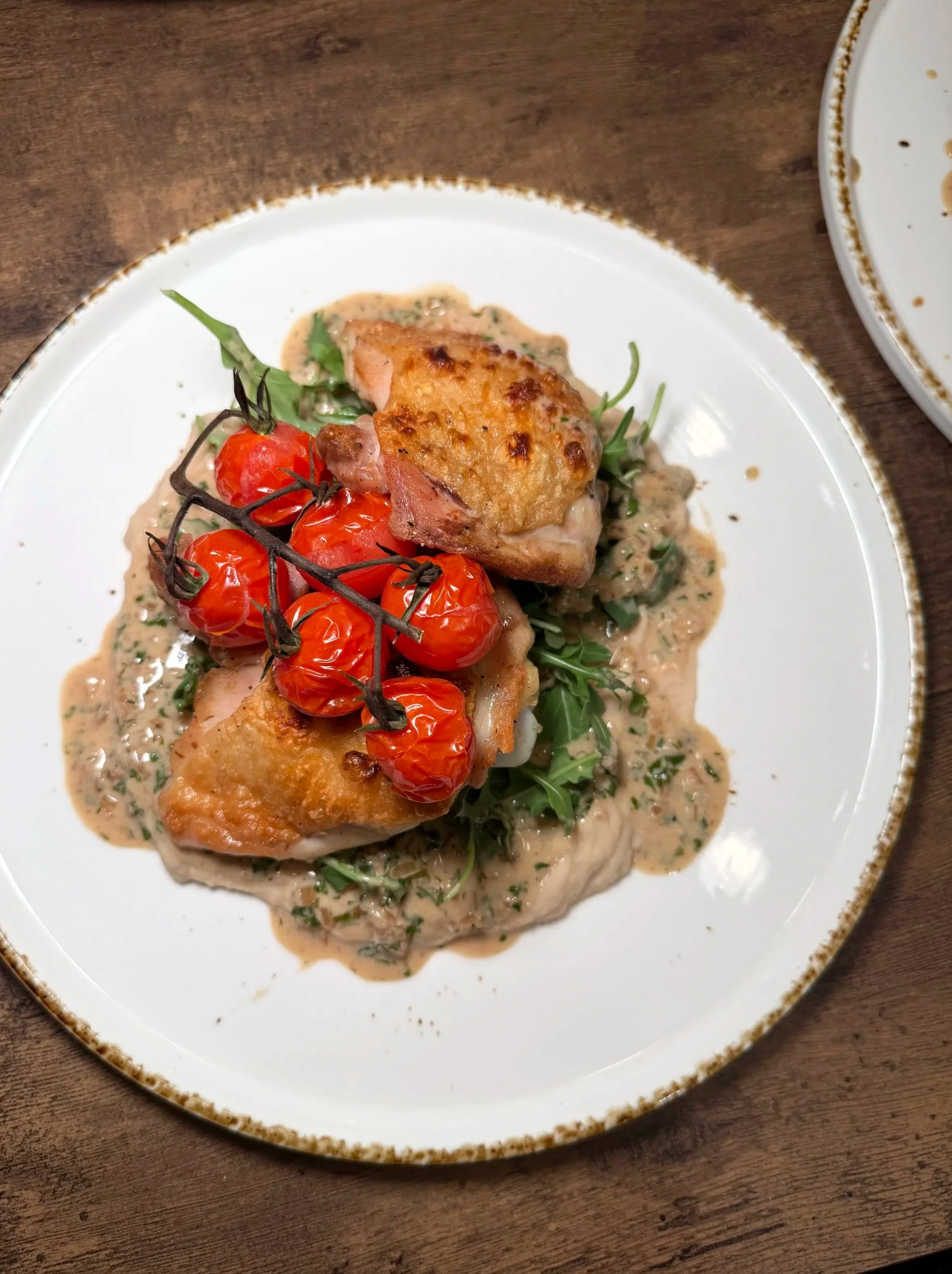 Plate of roasted chicken with cherry tomatoes on a bed of greens and creamy sauce on a wooden table.