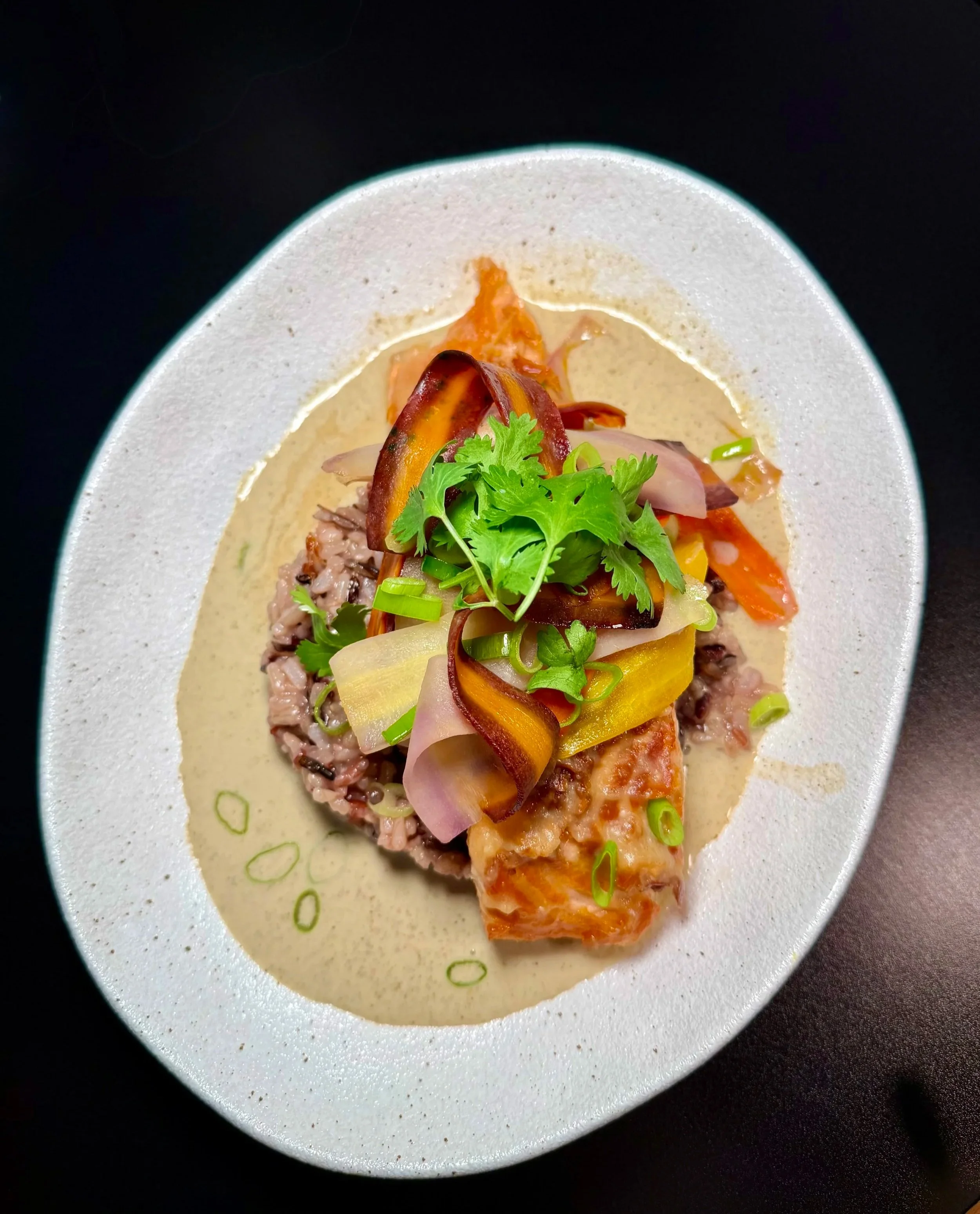 Close-up of a dish with rice, grilled chicken, roasted vegetables, and garnished with cilantro and green onions, served on a white plate with a black background.