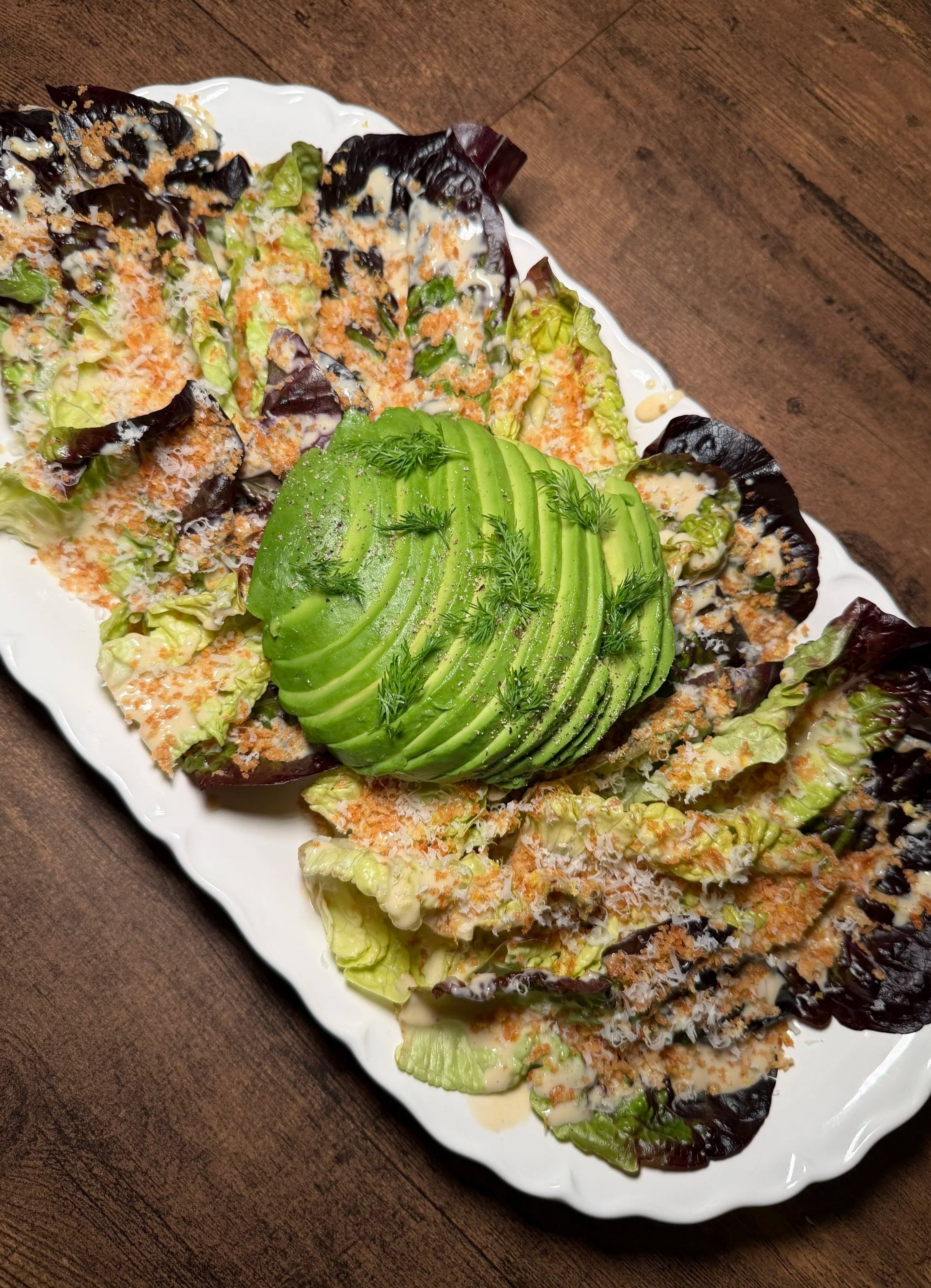 Salad with lettuce, sliced avocado, dill, and a creamy dressing, served on a white platter on a brown wooden table.