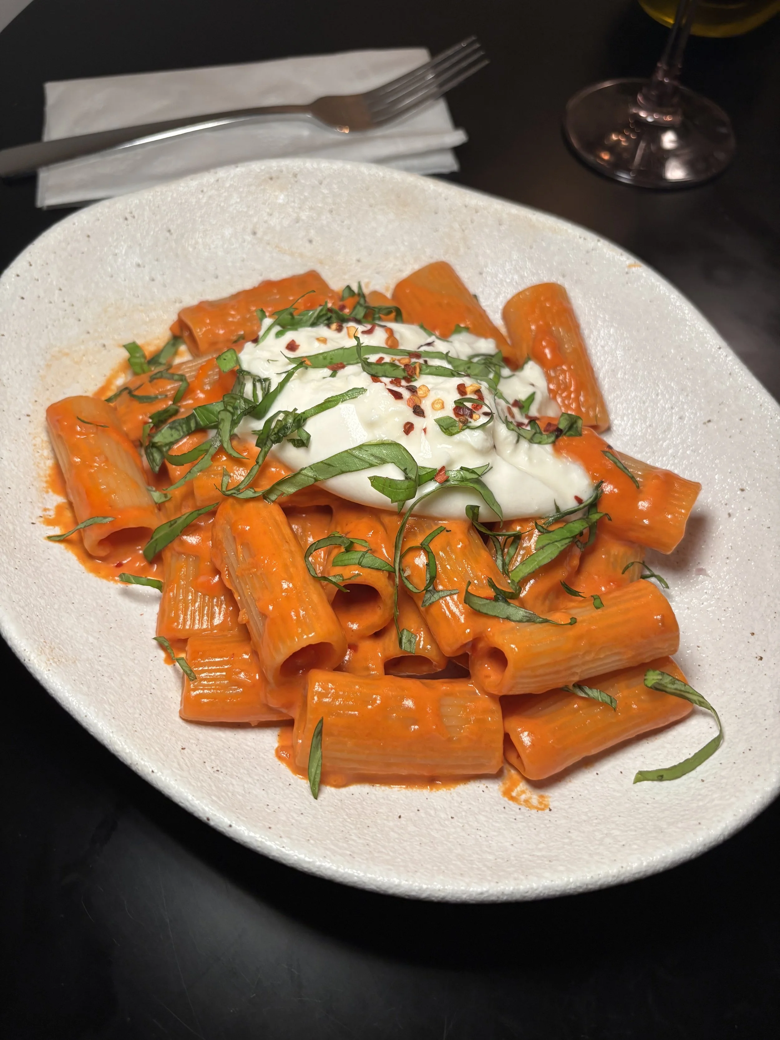 A white plate of cooked rigatoni pasta with tomato sauce, topped with sliced basil and red pepper flakes, on a black surface, with a fork, napkin, and wine glass nearby.