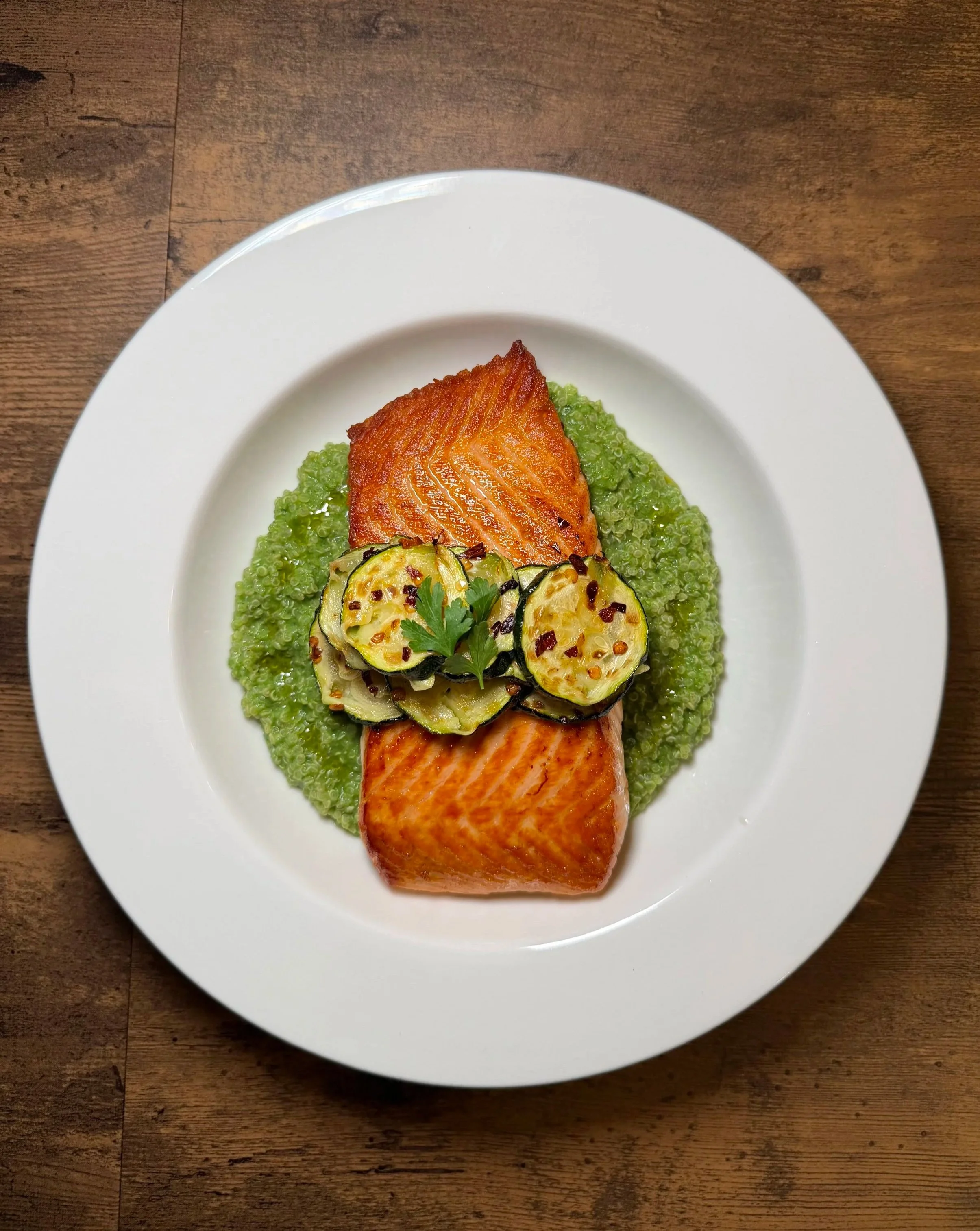 A white plate with cooked salmon fillet, grilled zucchini slices garnished with parsley and chili flakes, set on a wooden table.
