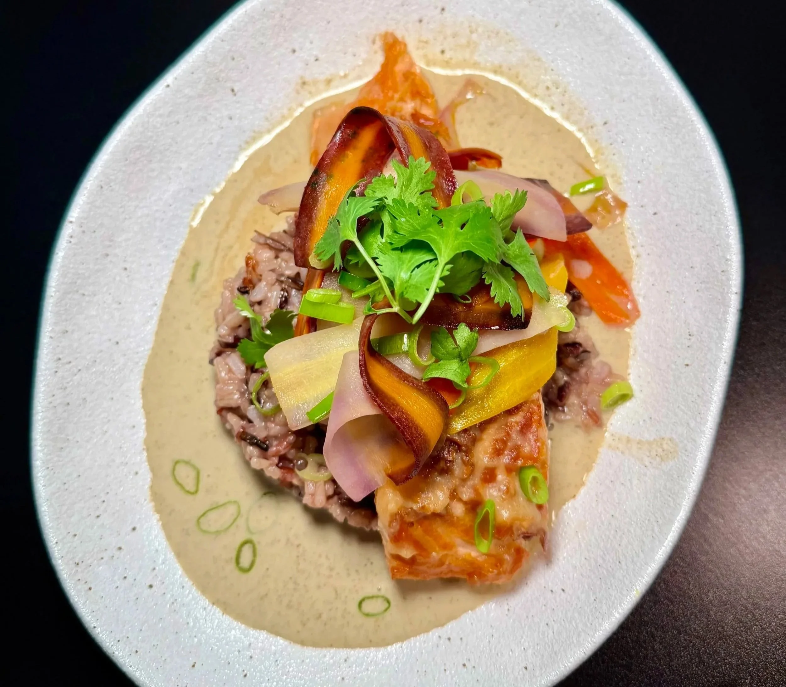 A plated dish featuring a piece of cooked fish topped with sautéed vegetables, fresh cilantro, and green onion, served with a creamy sauce.