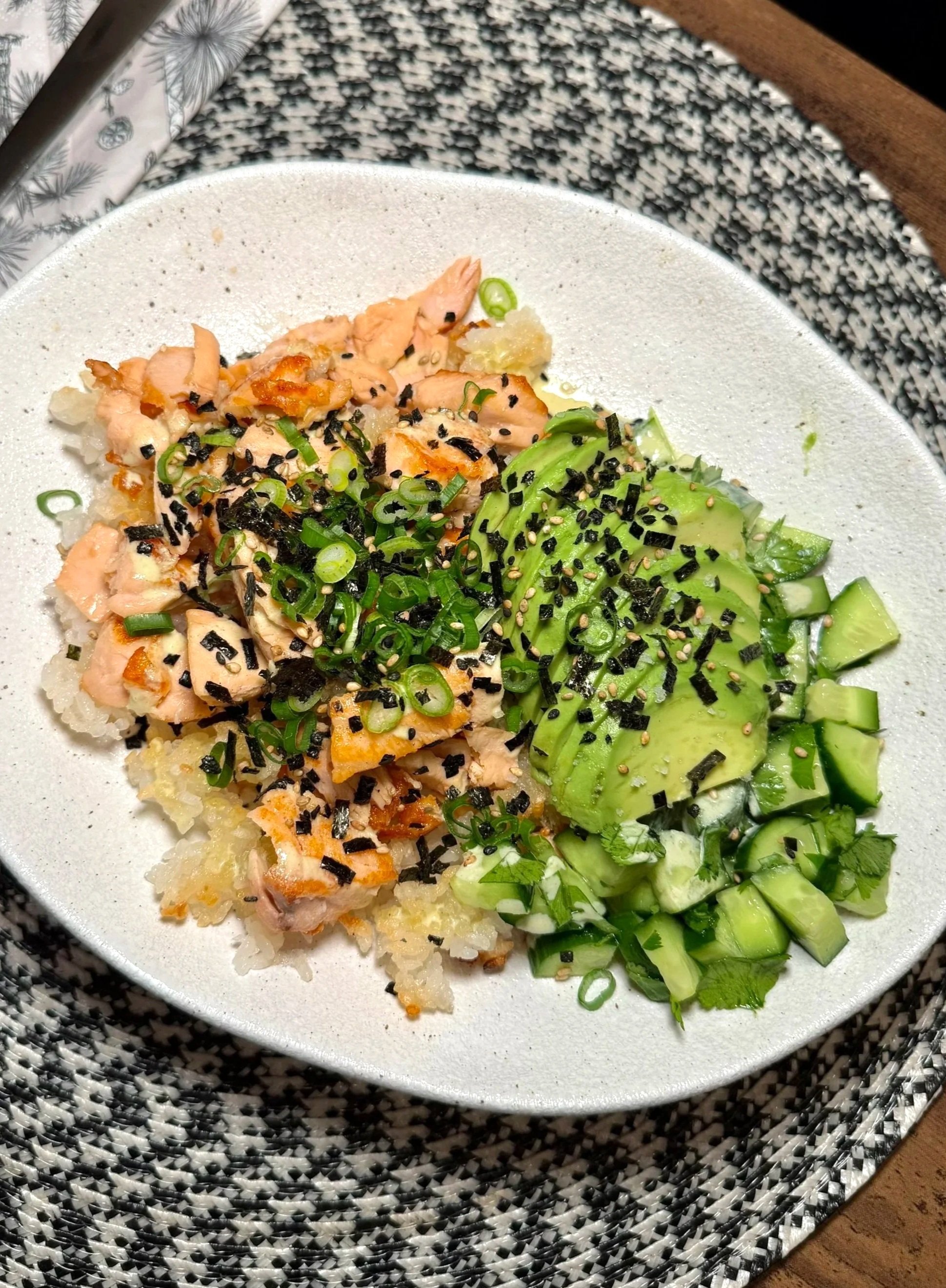 A white plate with sushi rolls topped with green onions, black seaweed, and sesame seeds, placed on a black and white woven placemat on a wooden table.