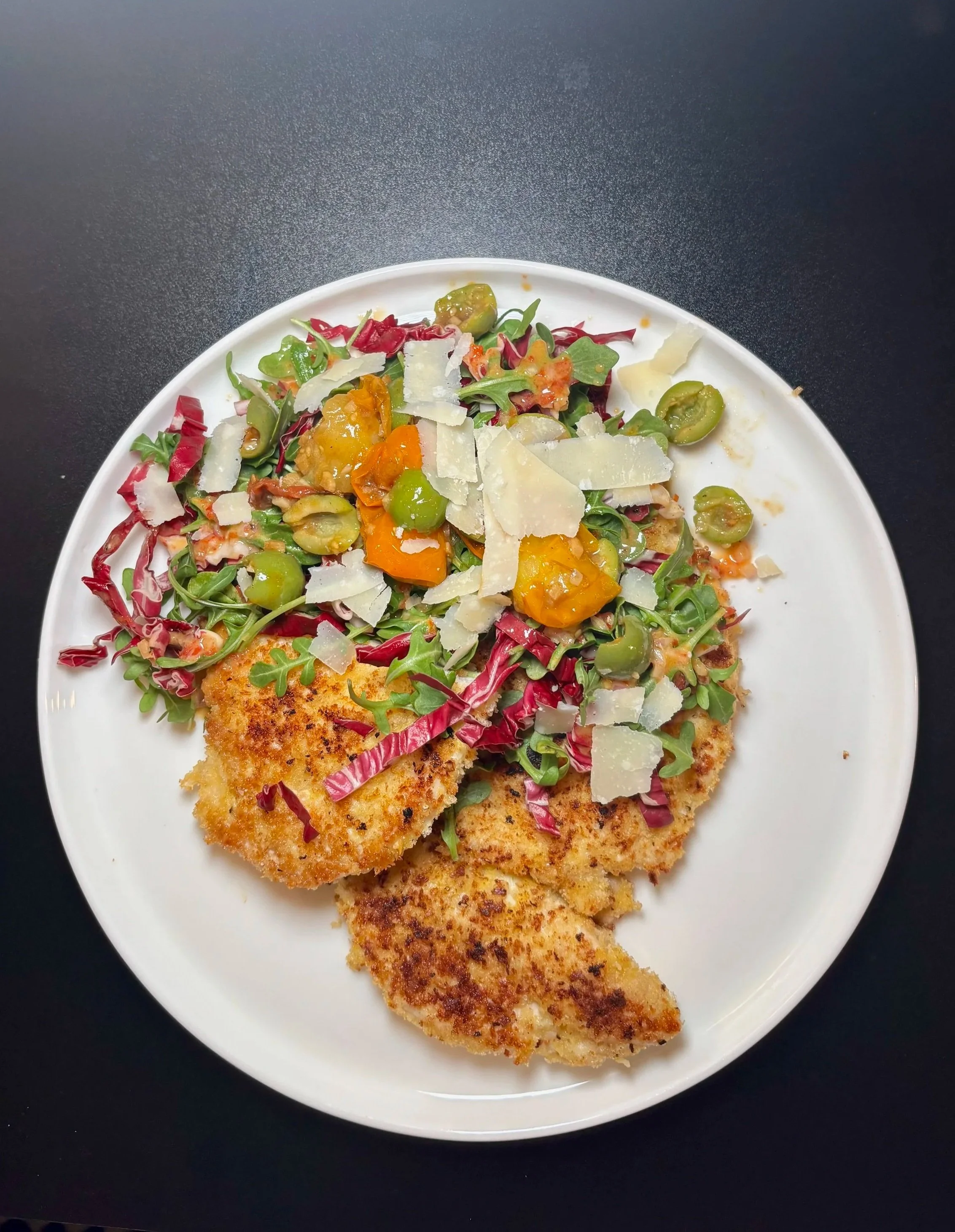 A white plate with breaded fish fillet, mixed green and red lettuce, sliced green olives, grated cheese, and tomato sauce, on a black surface.
