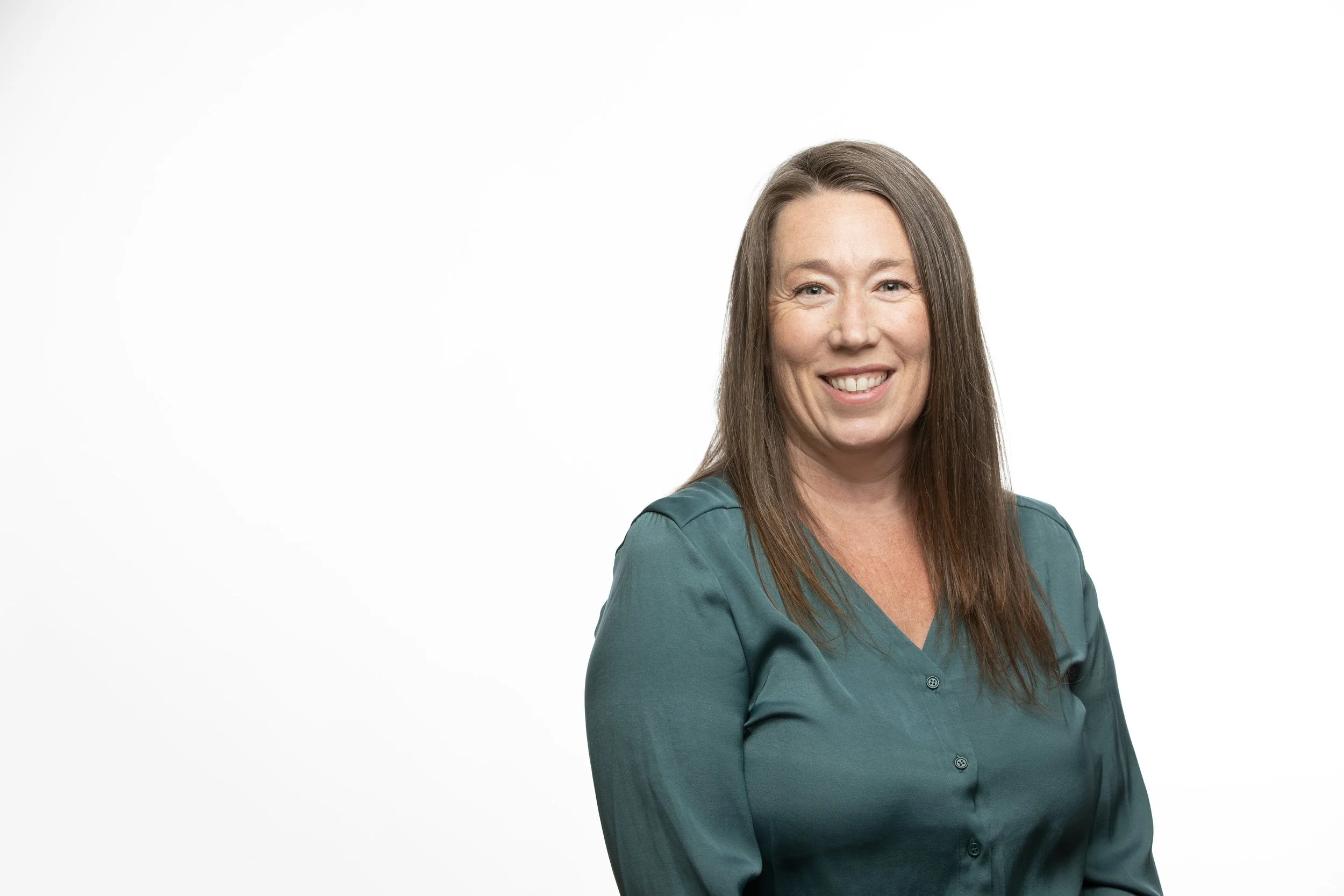 A woman with long brown hair, wearing a teal blouse, smiling, standing against a plain white background.