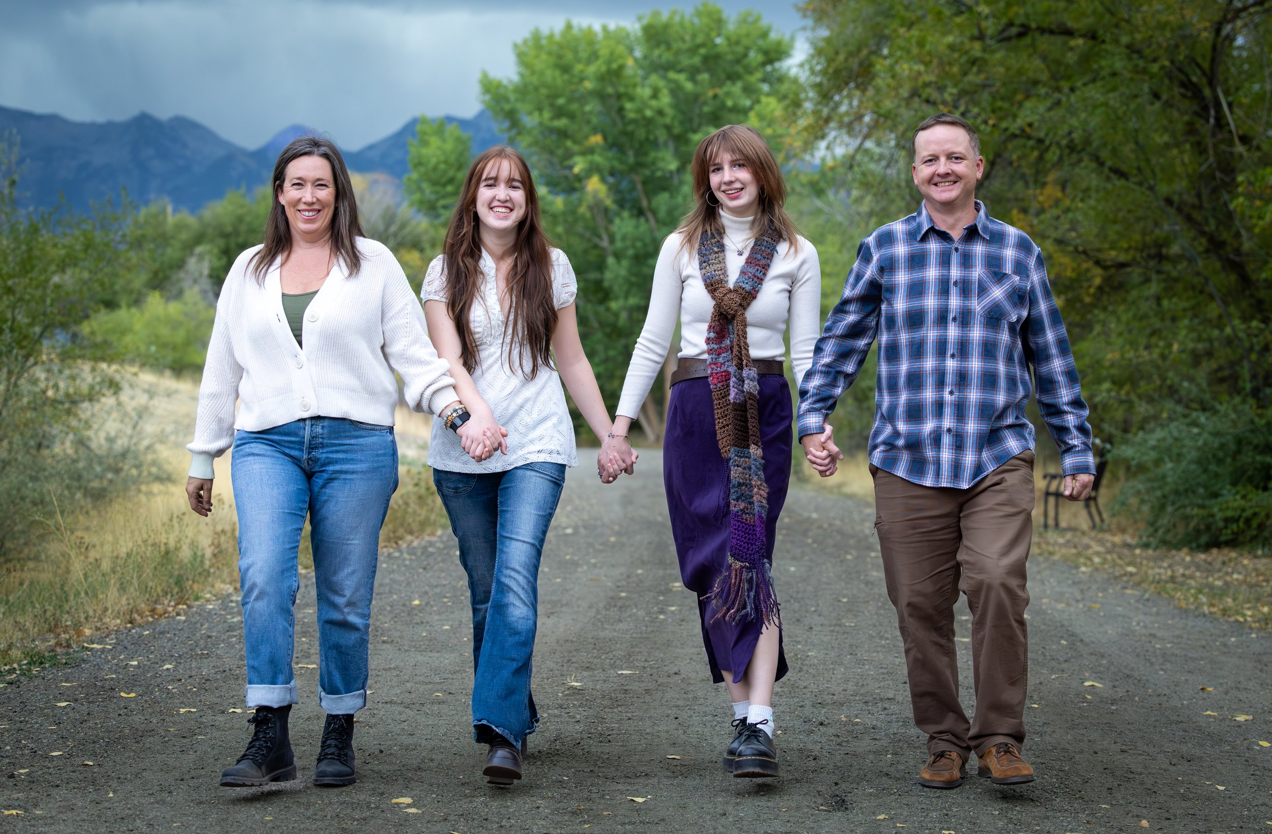 A family of four walking hand-in-hand outdoors on a trail with lush green trees and mountains in the background.