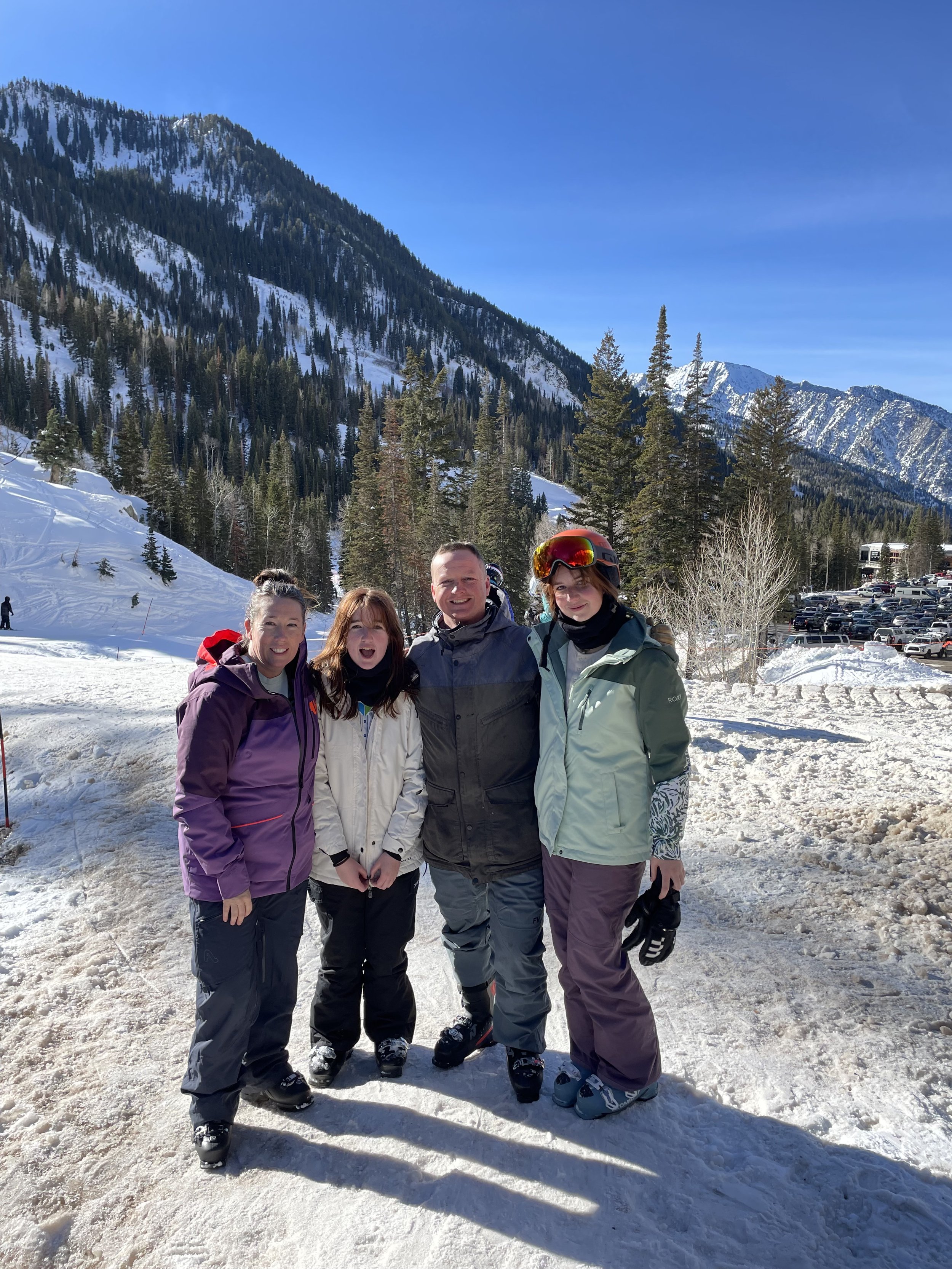 Four people in winter clothing standing on snow in a mountainous area with pine trees and snow-covered peaks in the background, smiling at the camera.
