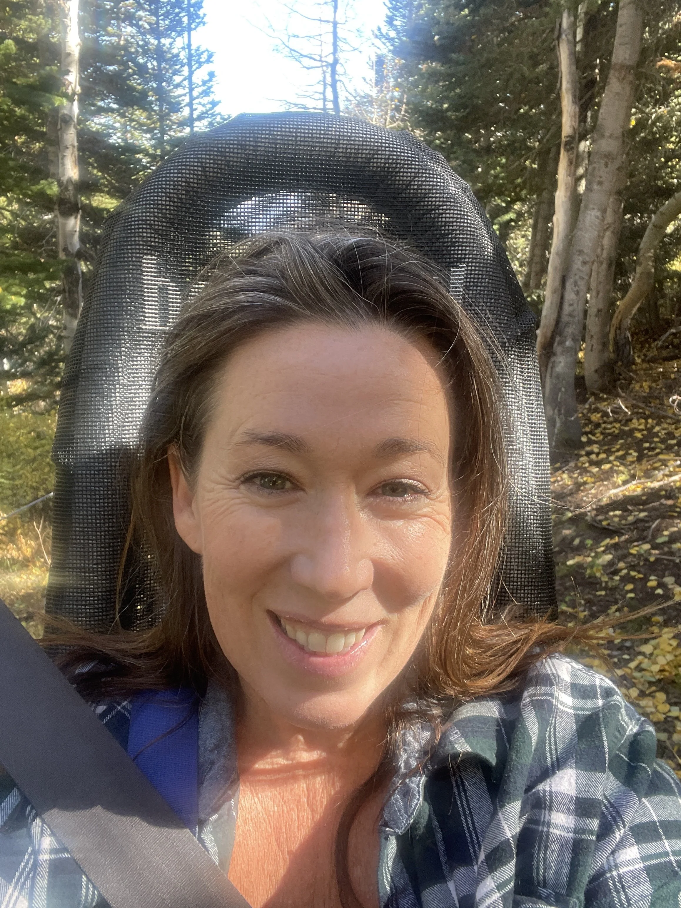 A woman smiling as she sits in a car, with trees and a clear blue sky visible outside.