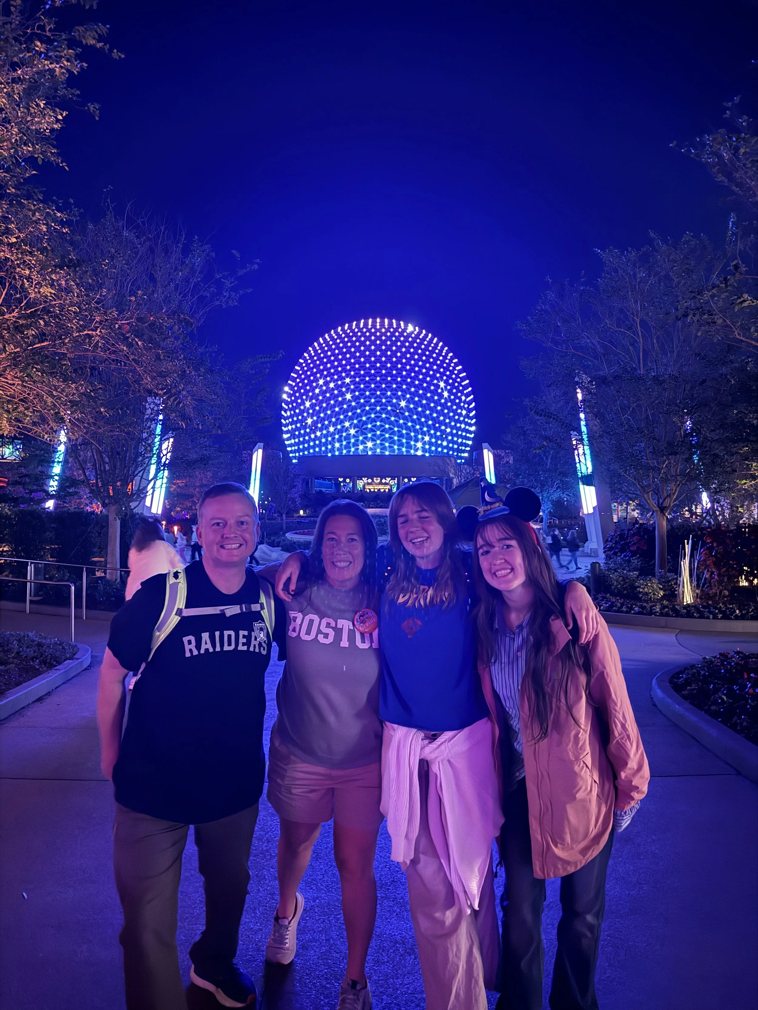 Four people smiling and standing close together at night outdoors, with a lit geodesic sphere structure and trees illuminated with blue and purple lights in the background.