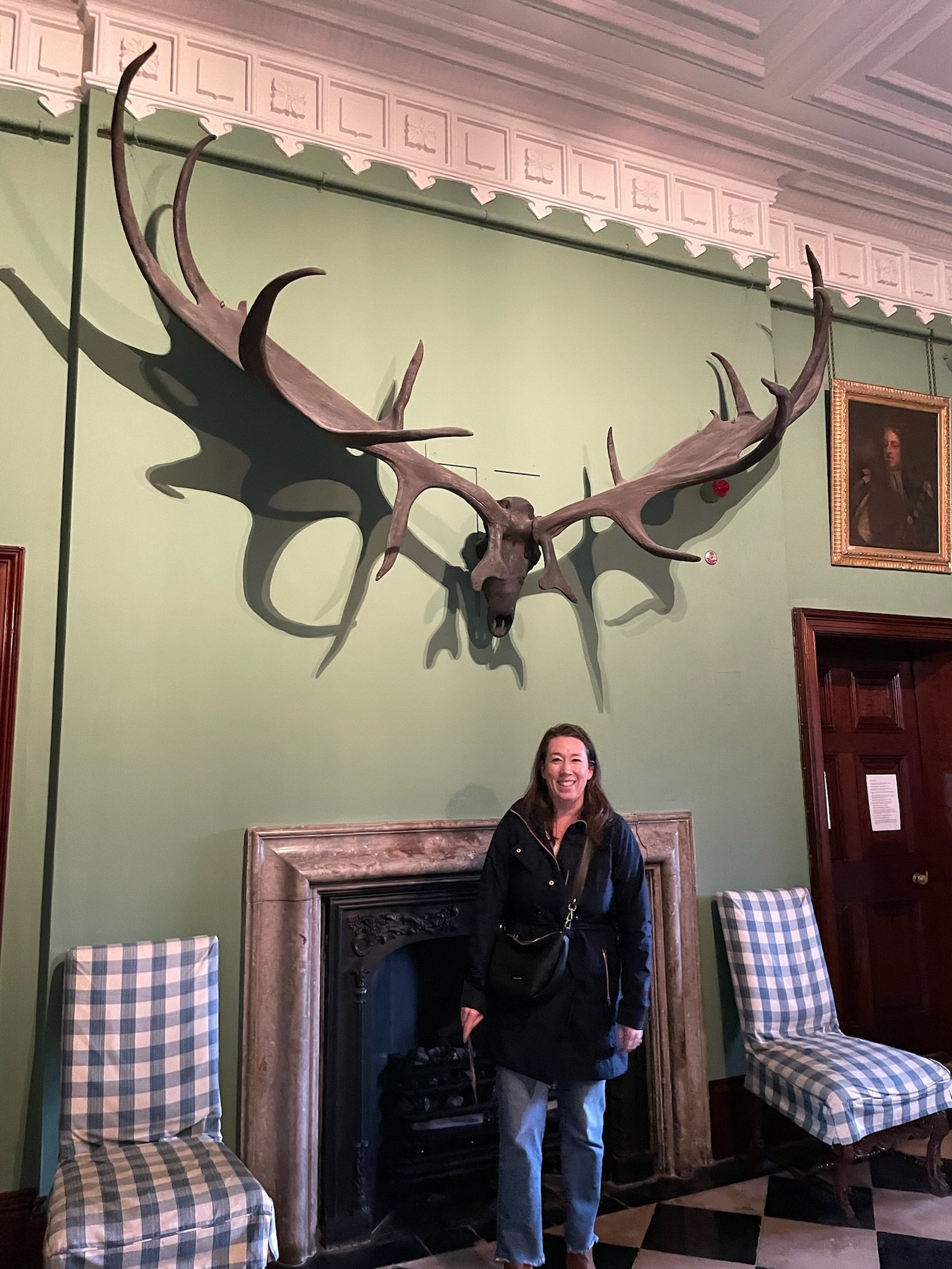 A woman with long hair smiling in a black jacket standing in front of a fireplace in a green-walled room. There are two striped armchairs with checkered fabric, one on each side of the fireplace. A large mounted antlered animal skull and a framed portrait are visible on the wall.