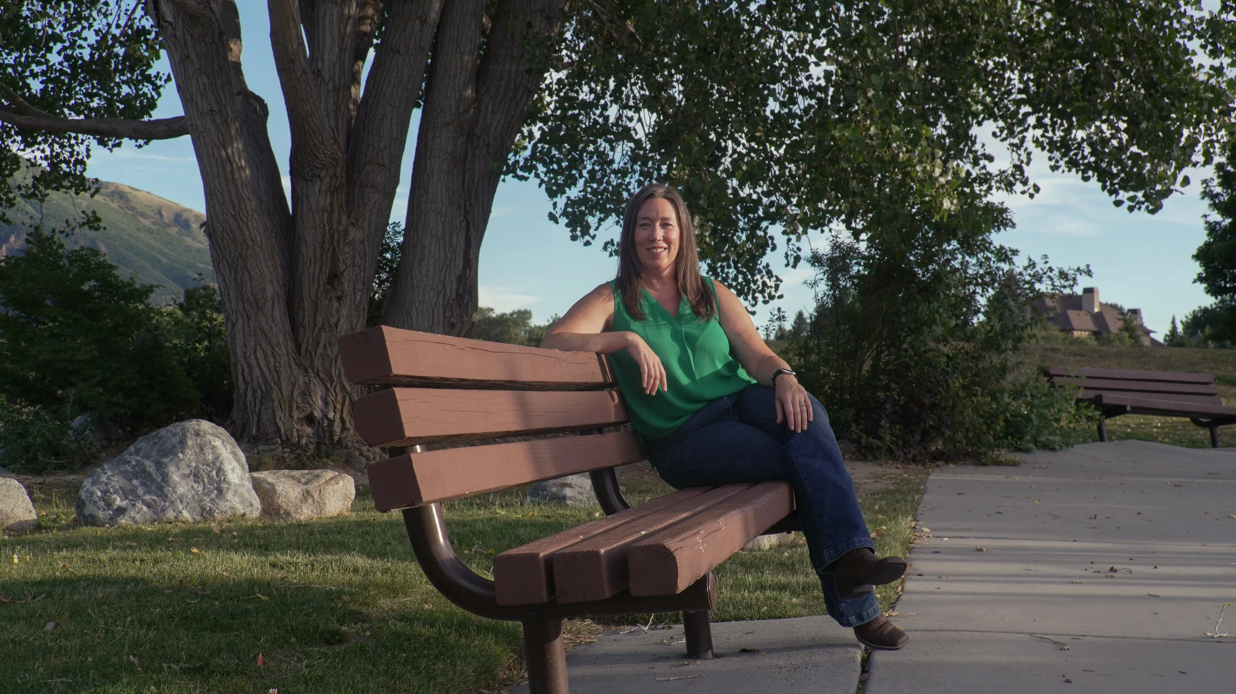 A woman in a green top and jeans sitting on a park bench beneath a large tree, smiling, with a mountainous landscape and houses in the background.