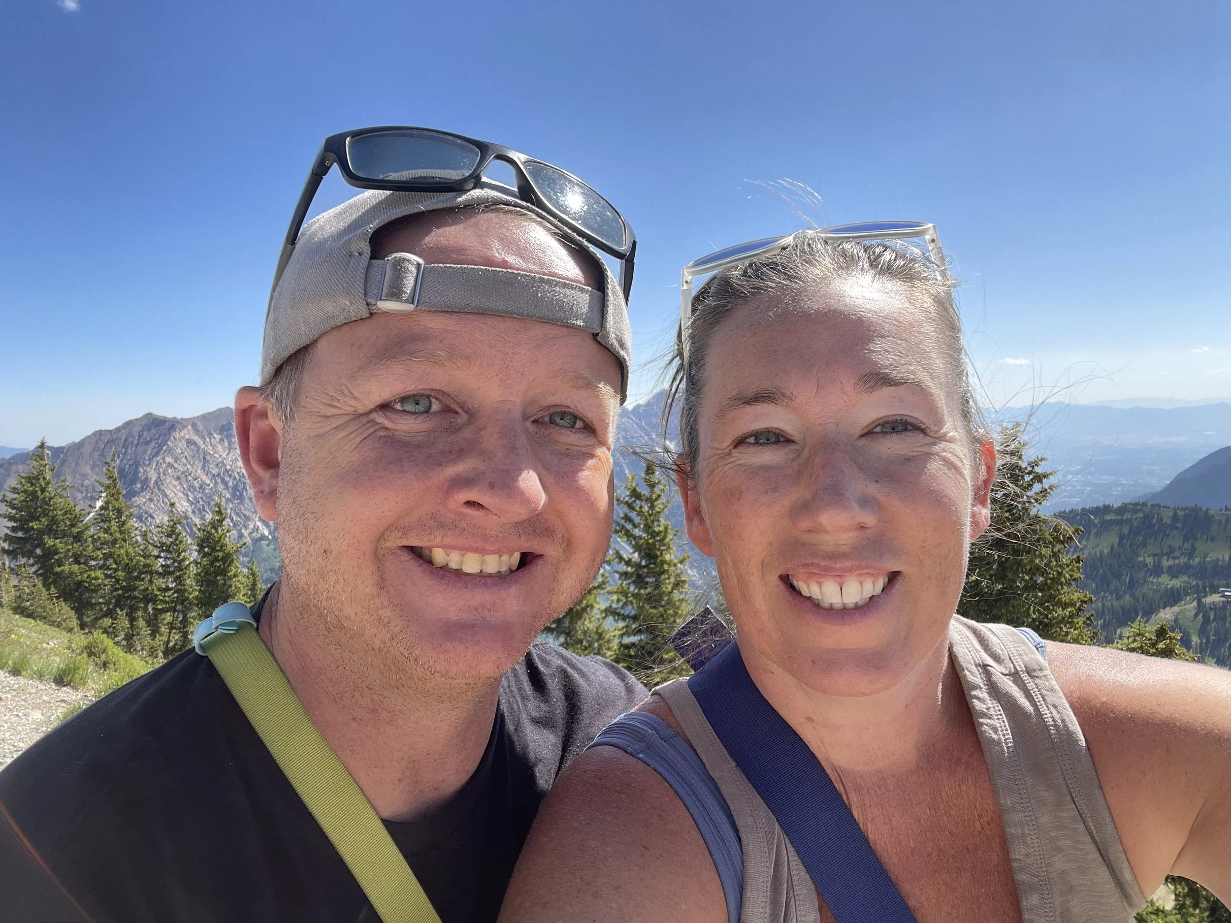 A smiling man and woman taking a selfie outdoors in a mountainous area under a clear blue sky, wearing casual hiking clothes and sunglasses.