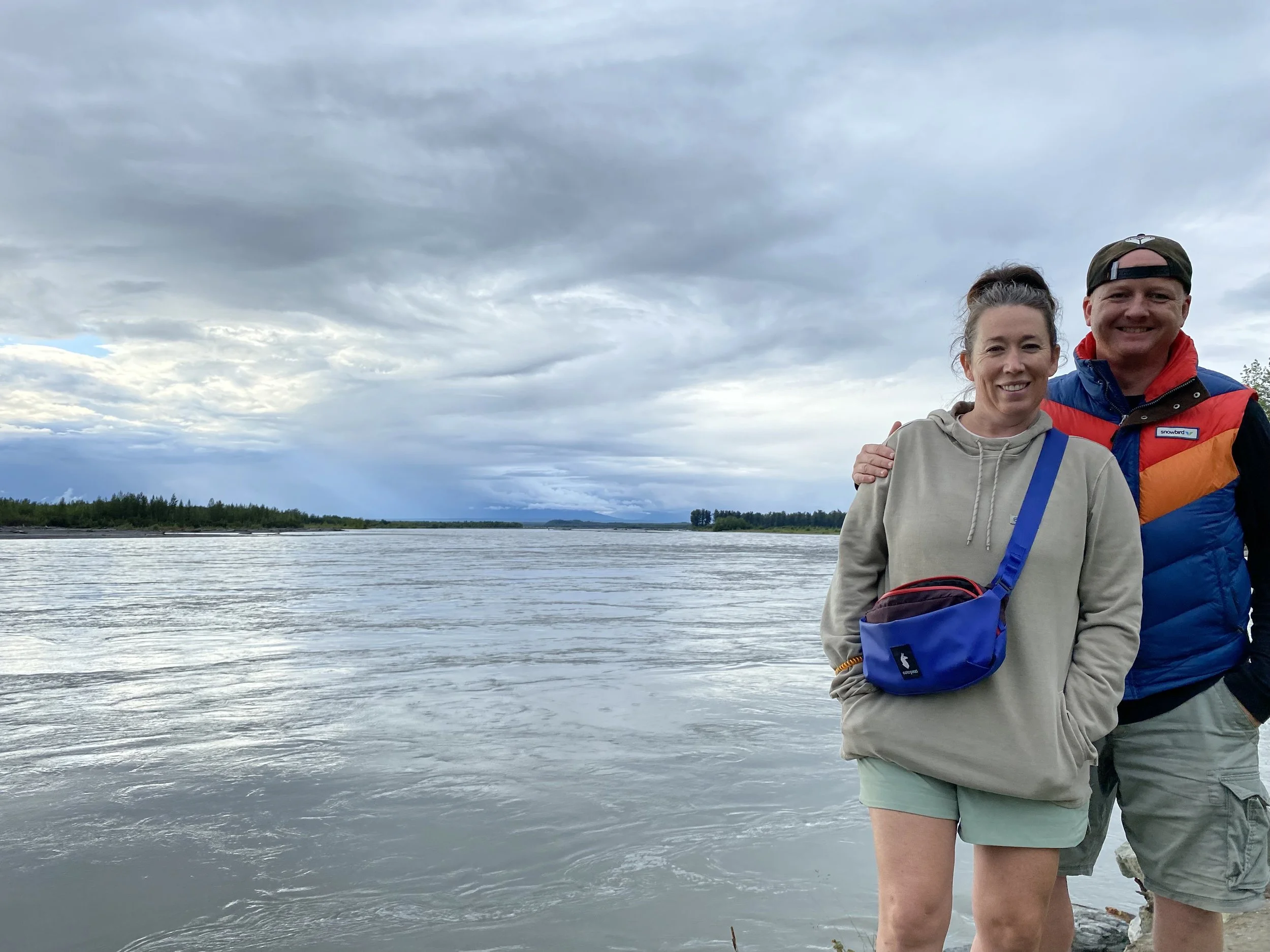 Two smiling people, a woman and a man, standing near the water with cloudy skies overhead.