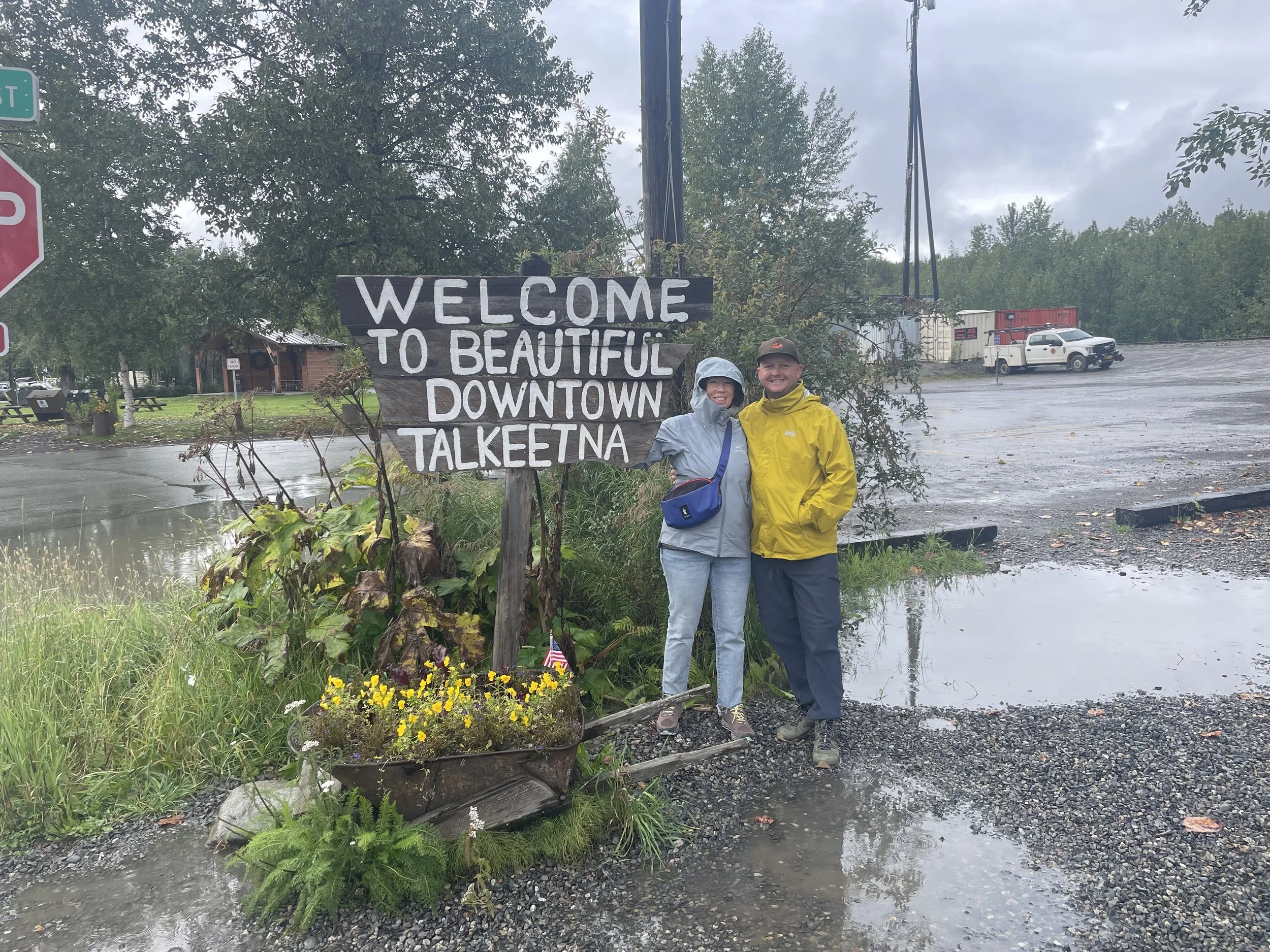 Two people standing outside on a rainy day in front of a wooden sign that reads 'Welcome to Beautiful Downtown Talkeetna,' with trees and parked vehicles in the background.