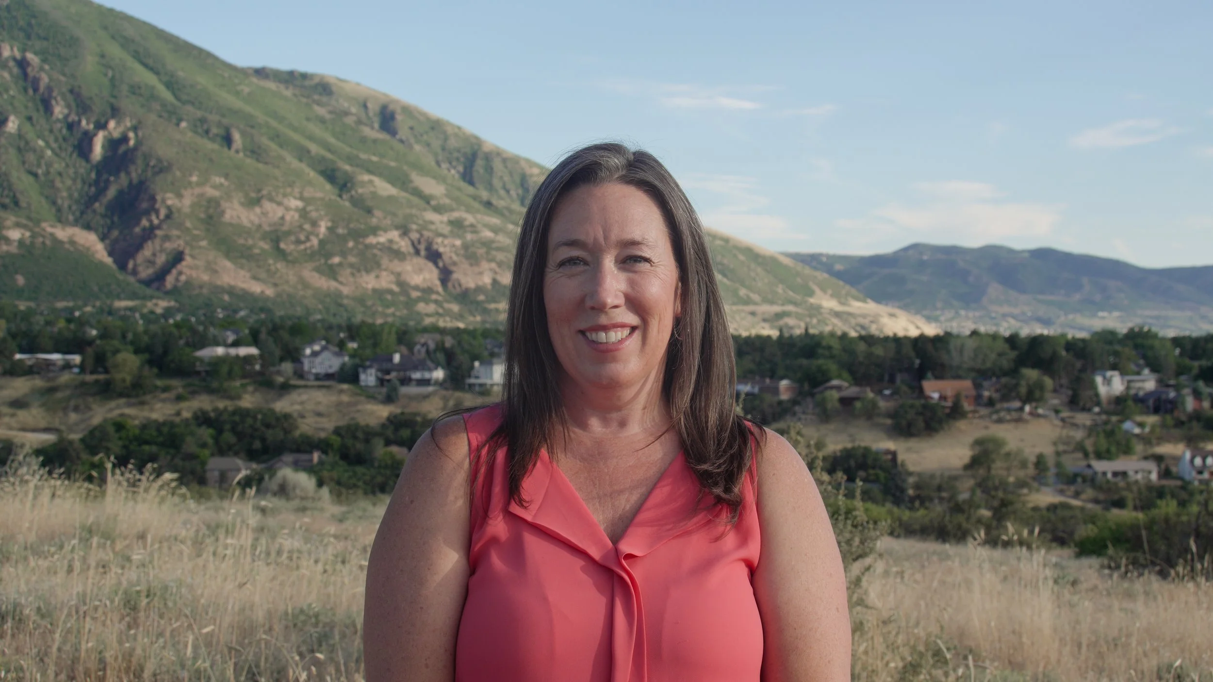 A woman with long dark hair wearing a pink sleeveless top, smiling outdoors, with green mountains, houses, and a blue sky in the background.