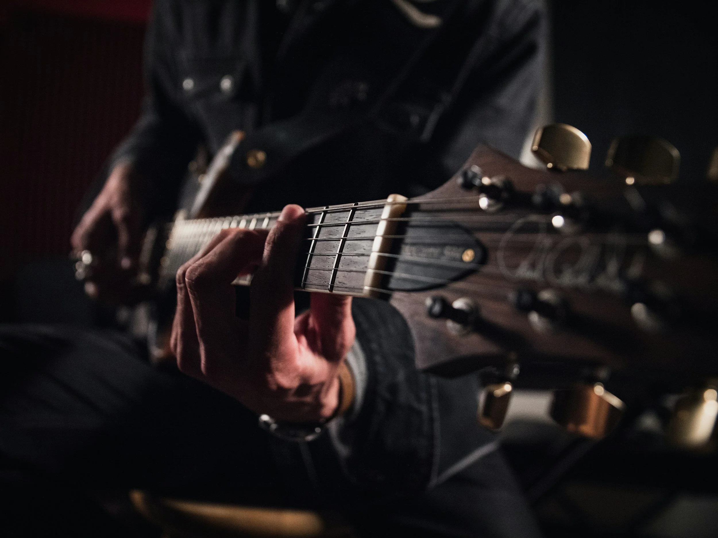 Close-up of a person playing an electric guitar, focusing on their hand on the fretboard, with dim lighting and blurred background.