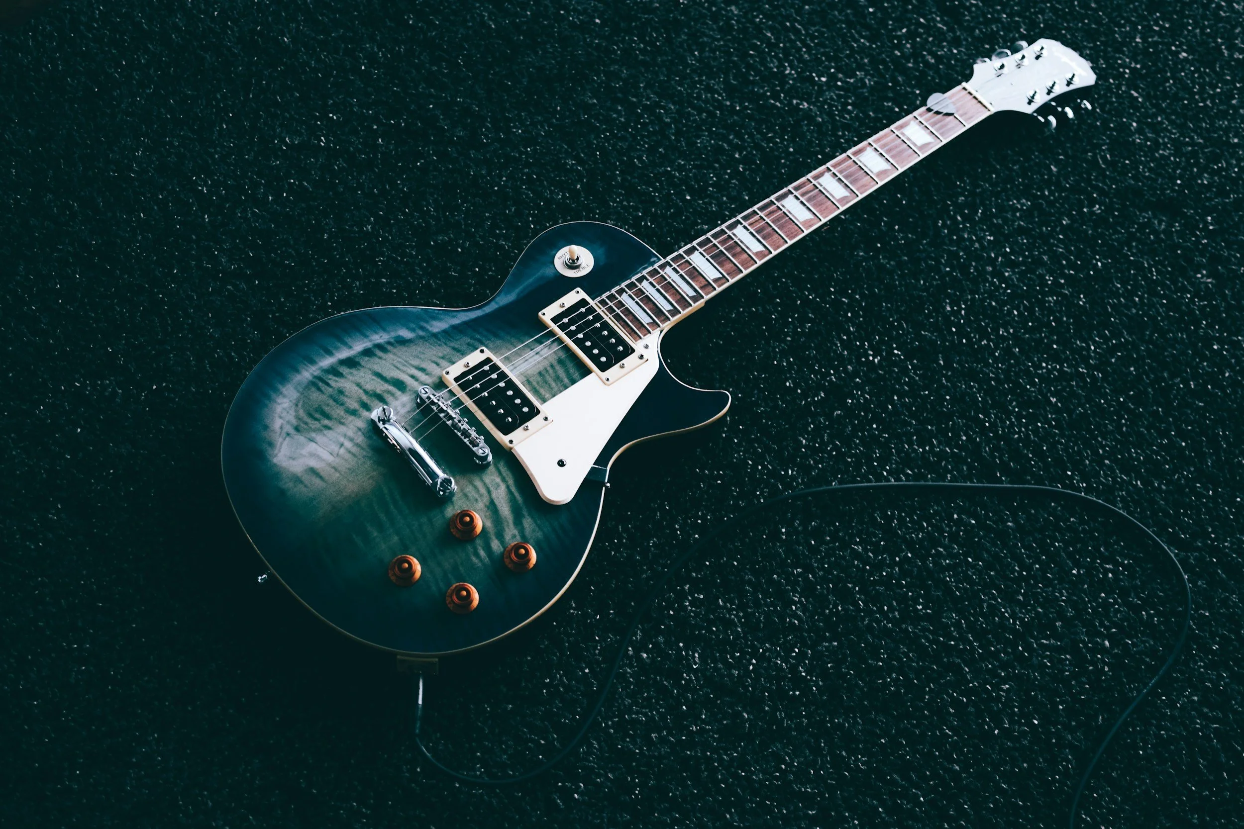 An electric guitar with a blue flame maple top, white pickguard, and chrome hardware, lying on a black textured surface.