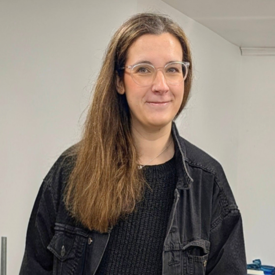 A young woman with long brown hair, wearing glasses, a black sweater, and a black denim jacket, standing indoors against a plain white wall.