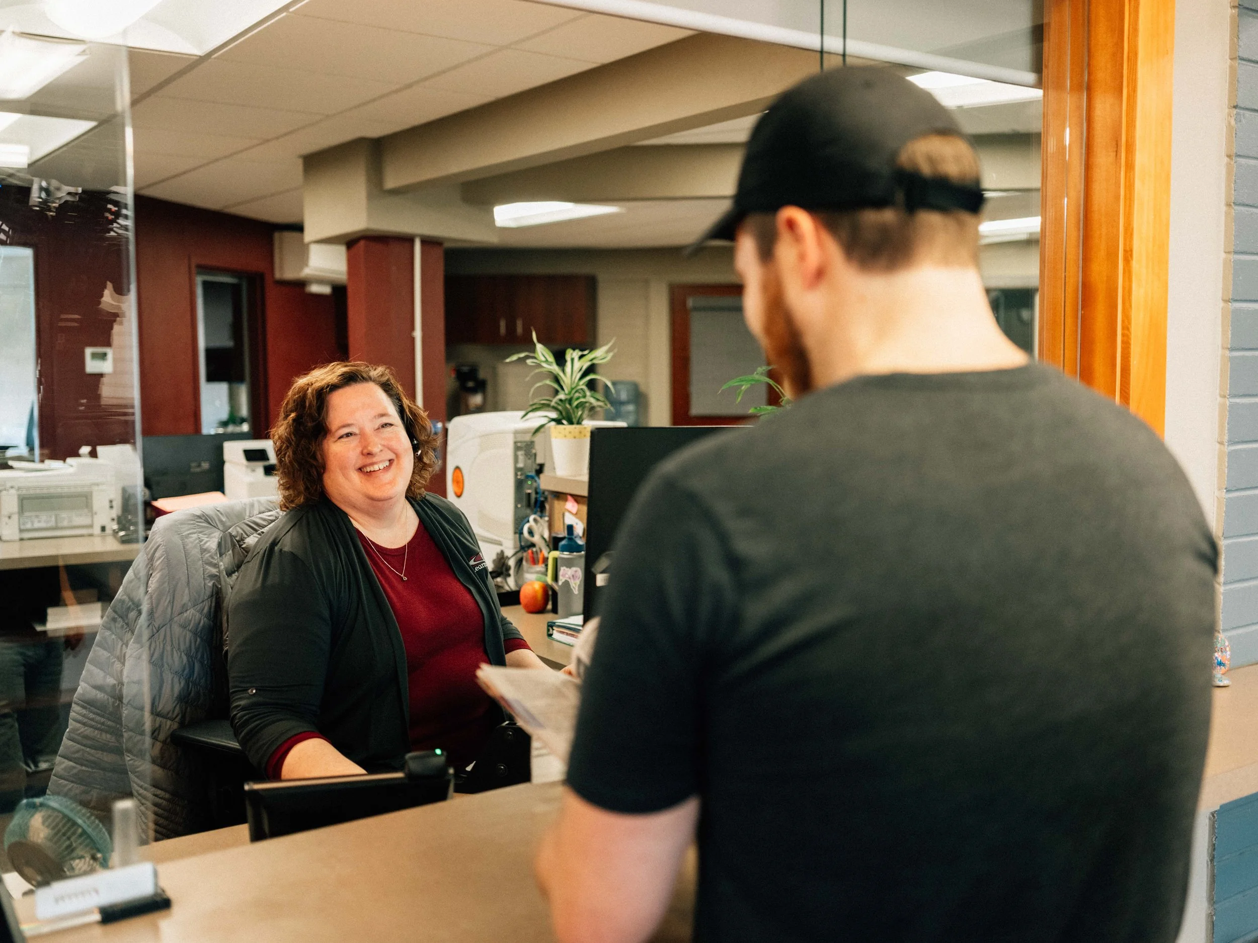 A woman smiling and seated at a reception desk, talking to a man in a black cap who is holding papers. The scene is in an indoor office environment with a plant, computer, and office supplies visible on the desk.