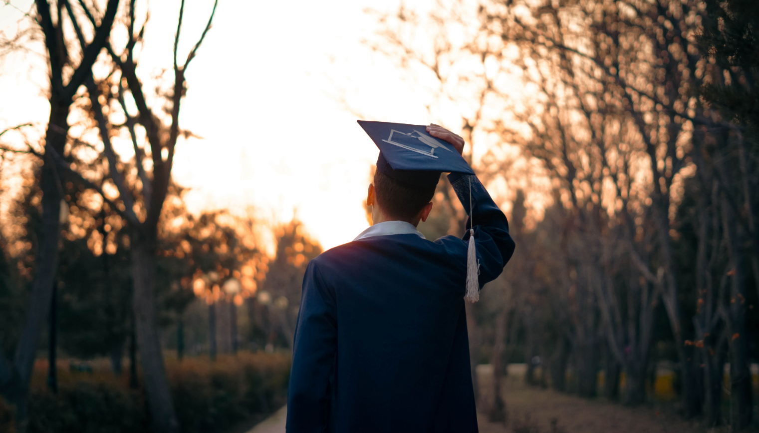 A person in a graduation gown holding their cap, standing outdoors at sunset in a wooded area.
