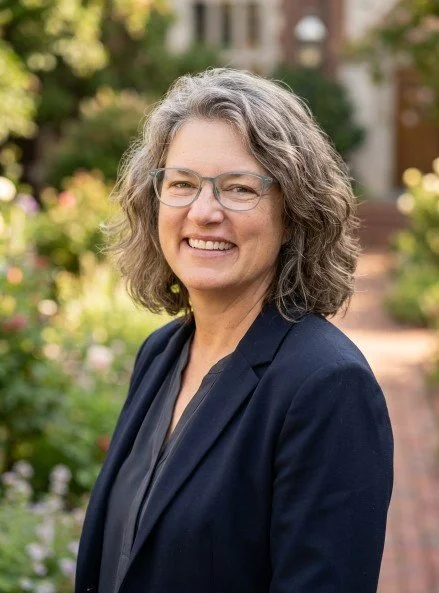 A smiling woman with gray curly hair and glasses, wearing a dark blazer, outdoors in a garden setting.