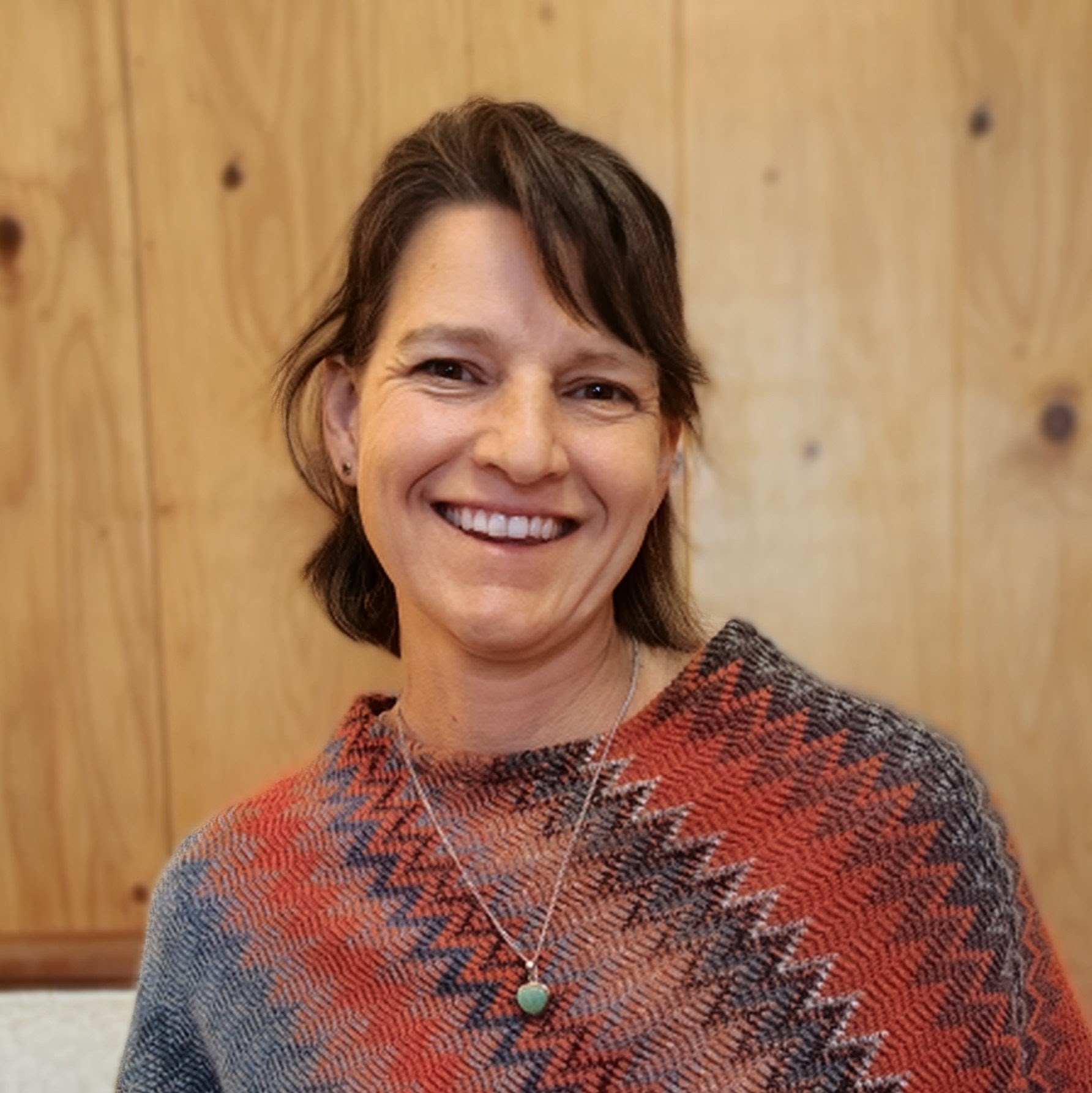 Close-up of a smiling woman with short brown hair, wearing a patterned sweater and a heart-shaped pendant necklace, standing against a wooden wall background.