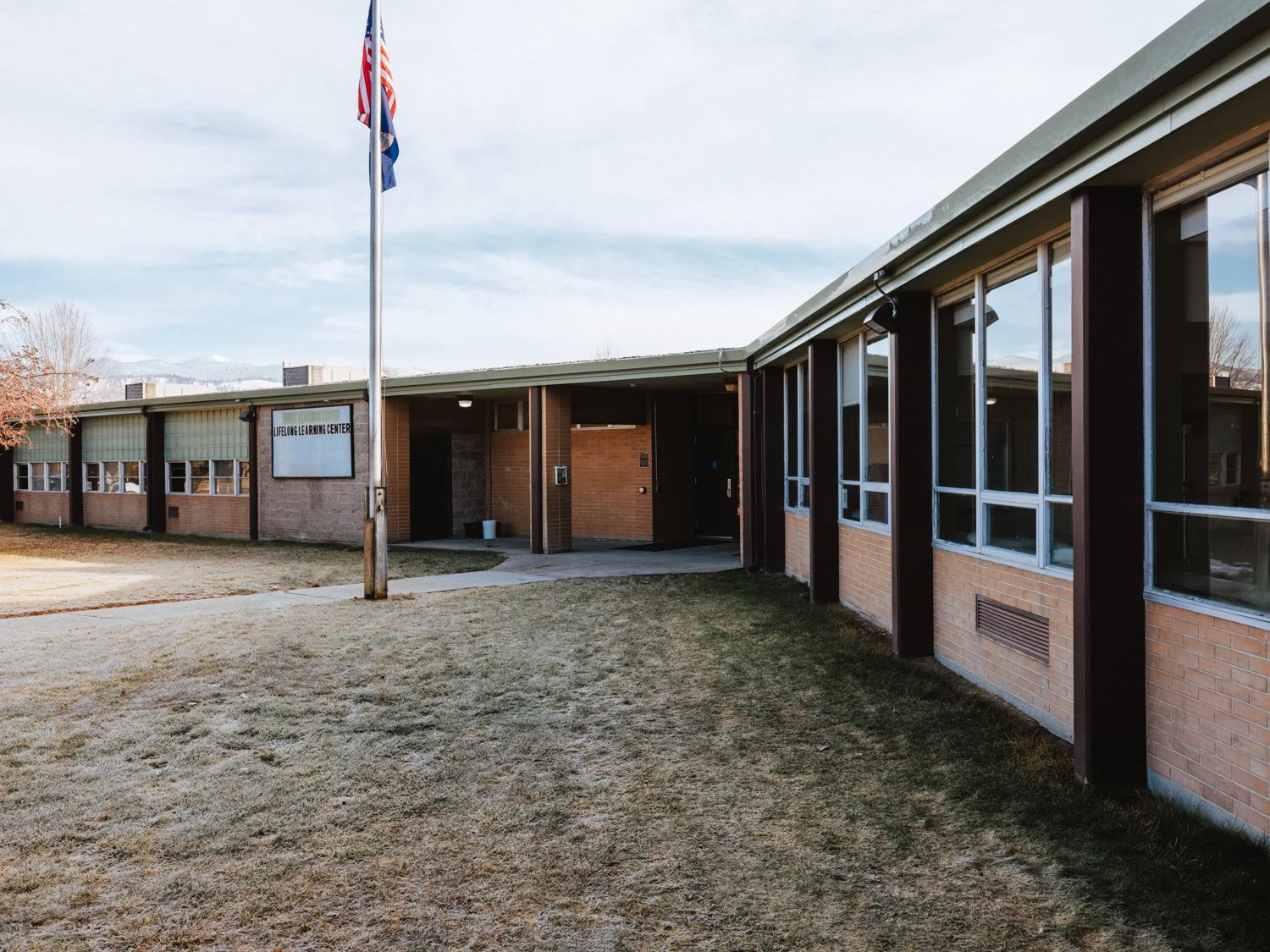A brick school building has a sign that reads 'Lifelong Learning Center' and a flagpole with an American flag in front of it, with a cloudy sky in the background and a lawn area in the foreground.
