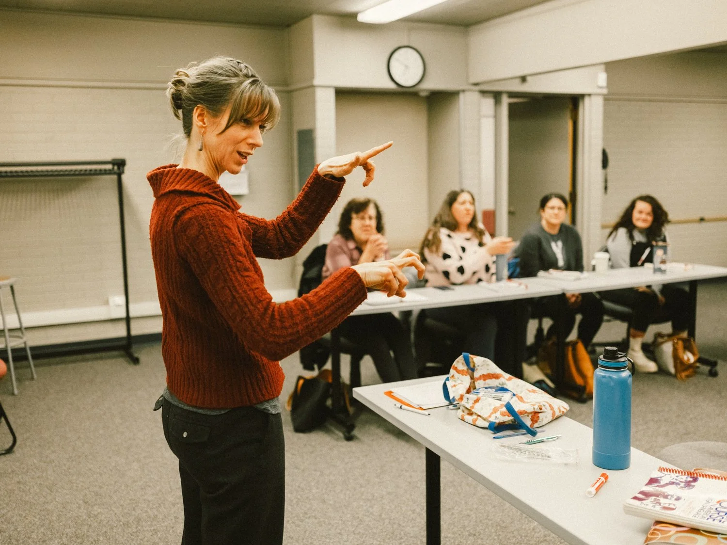 A woman with a red sweater standing and speaking to a group of four women seated at a table in a classroom or meeting room.