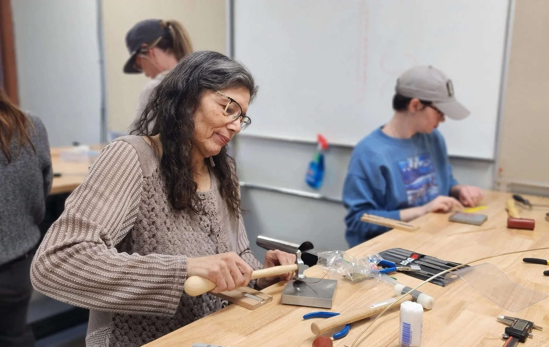 Woman working on a project at a woodworking workbench in a classroom with tools and supplies, while others work nearby.