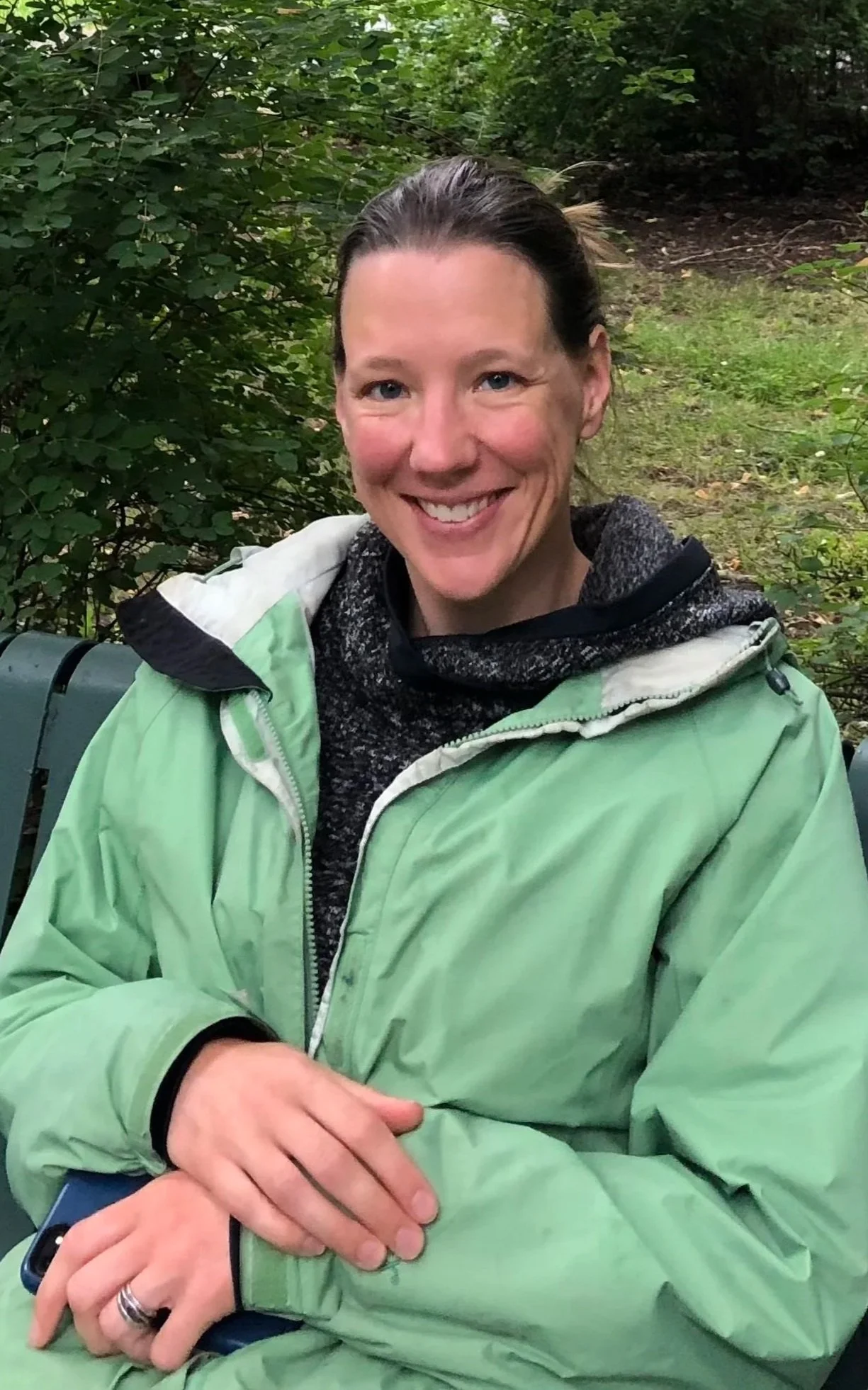 A woman smiling outdoors, wearing a green jacket and black hoodie, with her hair tied back, surrounded by green foliage.