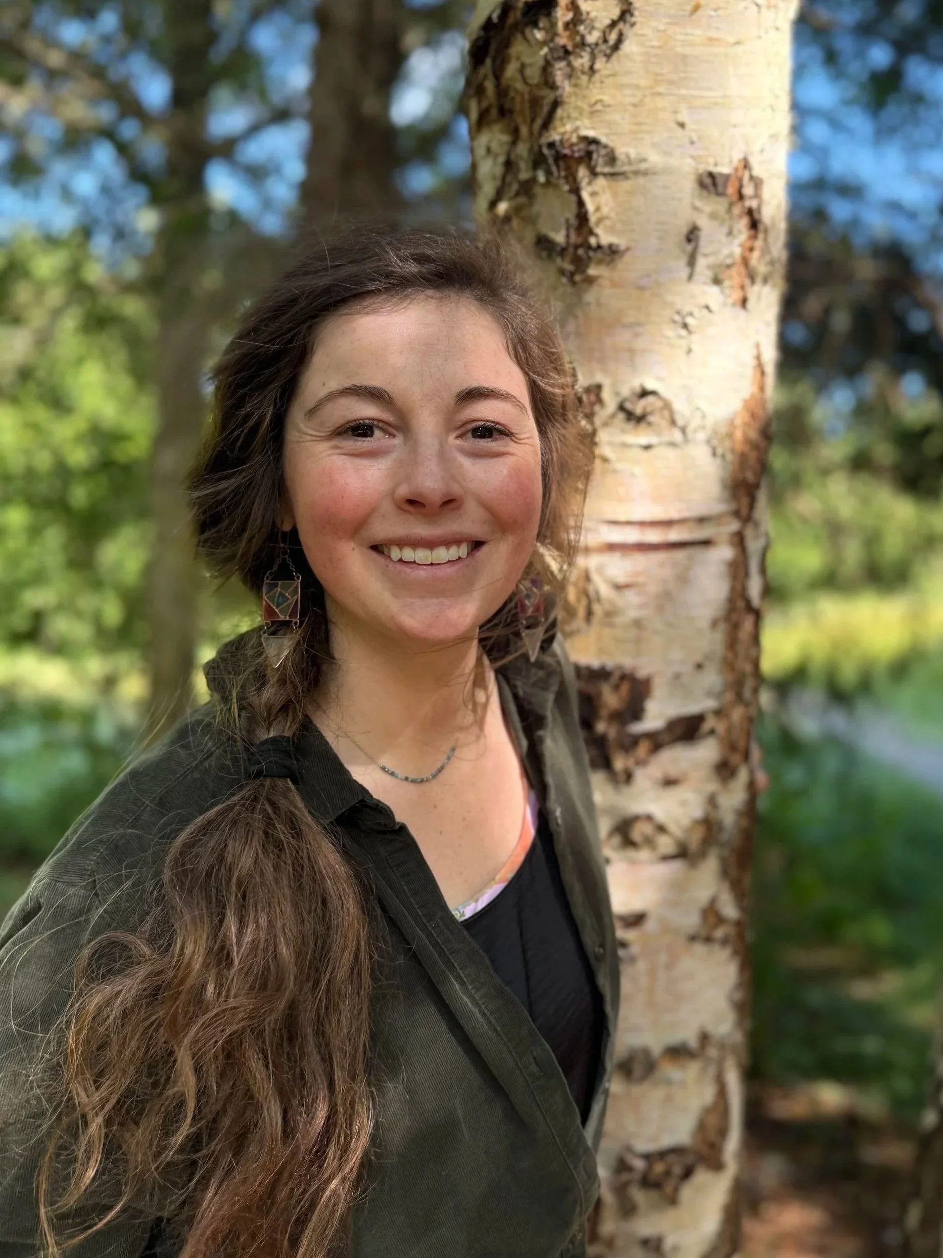 A woman with long brown hair and earrings smiling outdoors near a tree trunk with green and blue background.