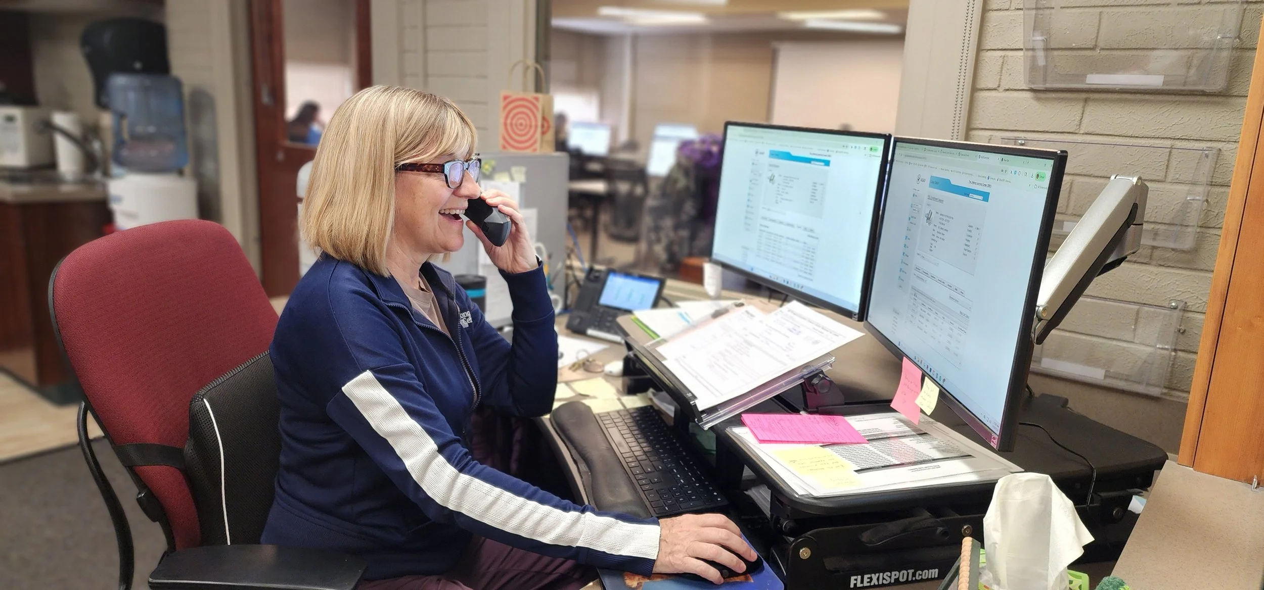 A woman with glasses, blonde hair, and a smile, is sitting at a cluttered office desk, talking on the phone, with dual computer monitors, documents, sticky notes, and office supplies.