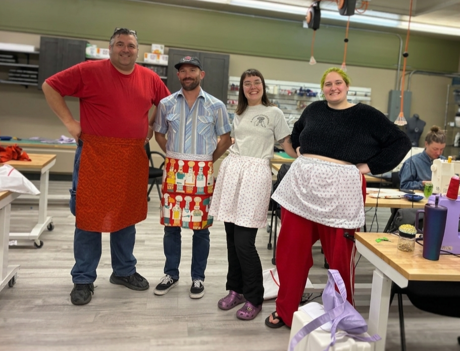 Four people standing in a room with sewing supplies, all wearing aprons, smiling at the camera.