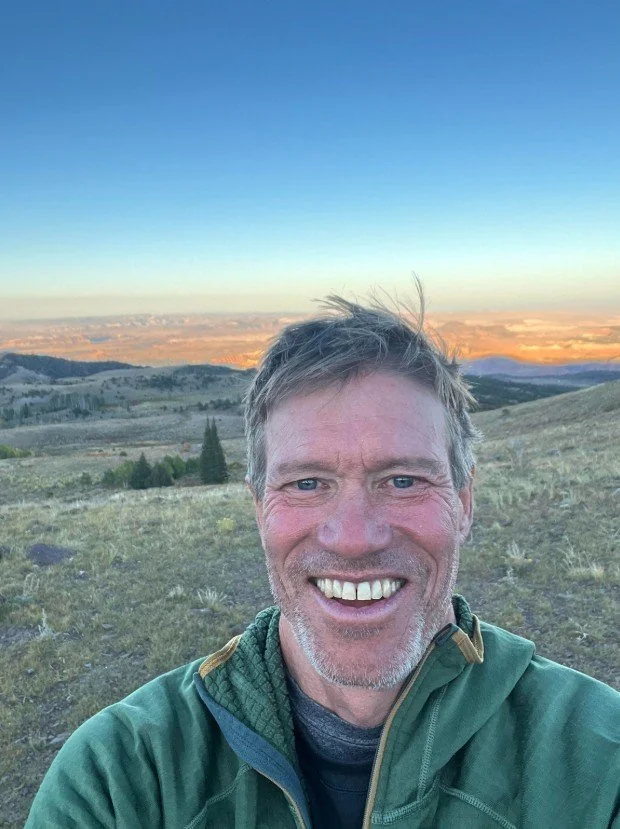 A smiling man taking a selfie outdoors on a hill with a scenic landscape and sunset in the background.