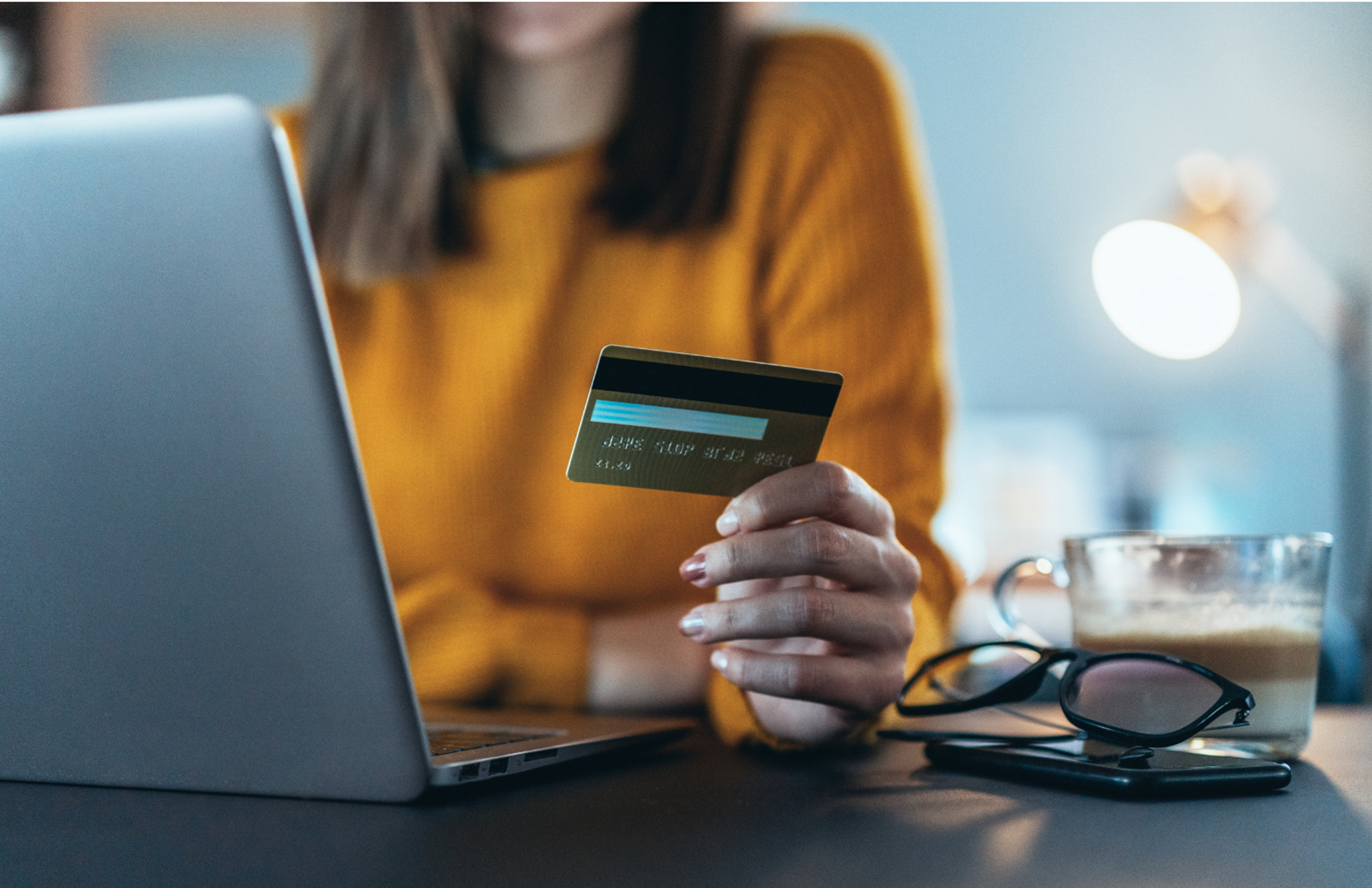 A woman in a mustard yellow sweater sitting at a desk, holding a credit card in her hand, with a laptop, glasses, a cup of coffee, and a pair of eyeglasses on the desk.