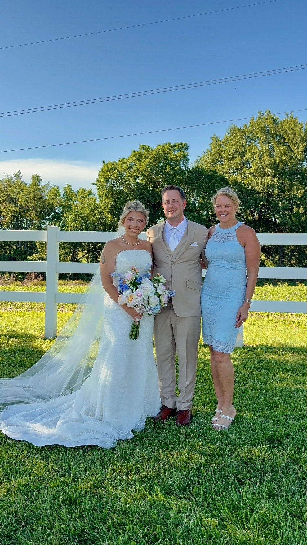 A bride and groom standing with a woman outdoors, with green trees and a white fence in the background, on a sunny day.